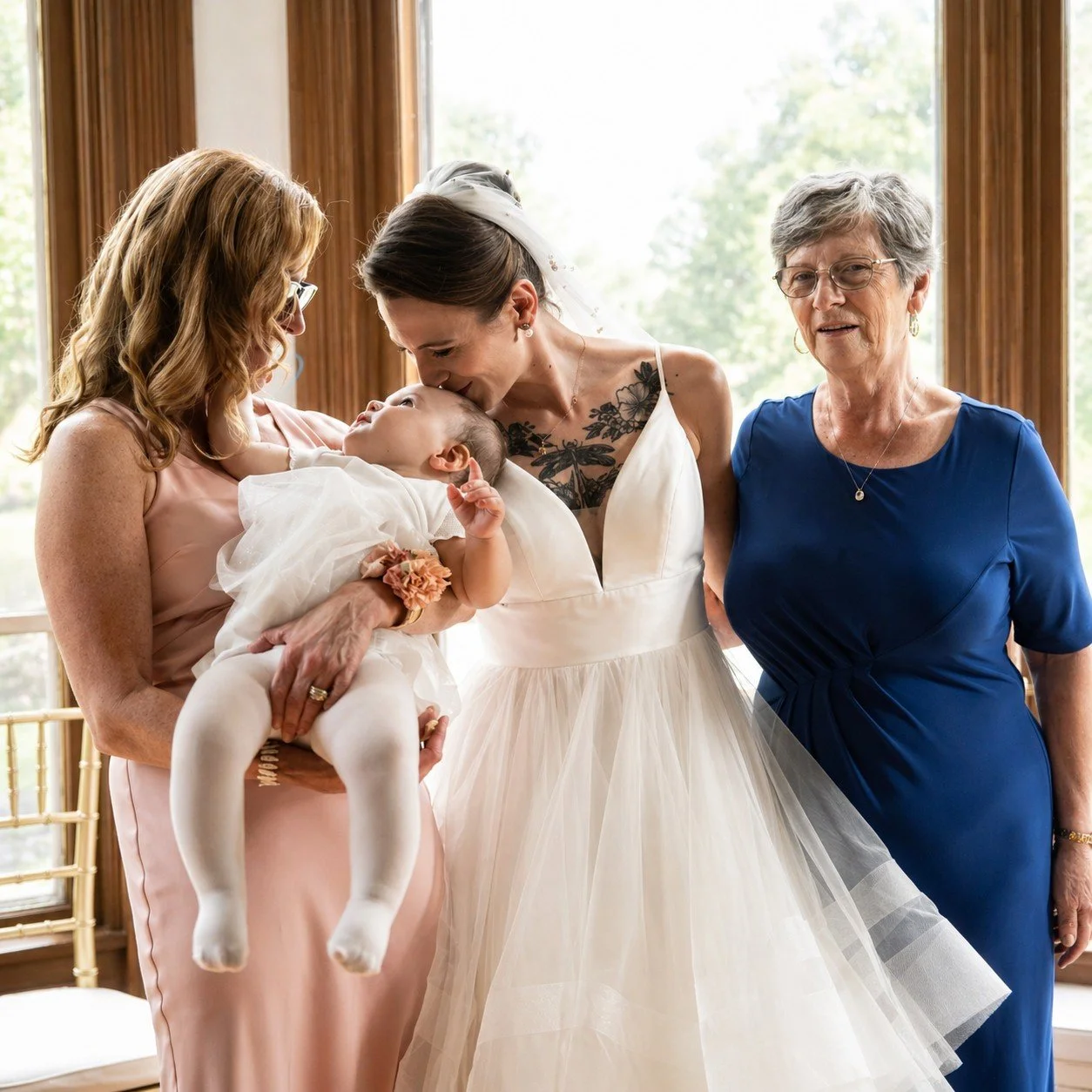 A bride, holding a baby, is kissing her mother on the forehead during a wedding celebration.