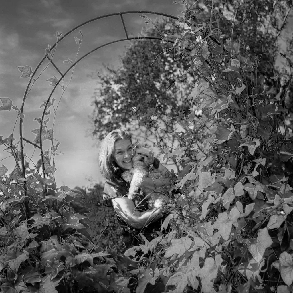 A woman hugging a small dog while standing among tall plants with a garden arch overhead.
