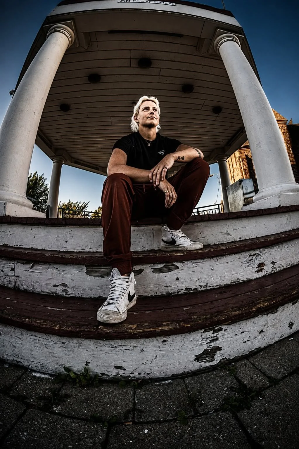 Woman with blonde hair sitting on the steps of a weathered white wooden porch with four large white columns, wearing a black t-shirt, brown pants, and white Nike sneakers with black swoosh, looking upward against a blue sky.