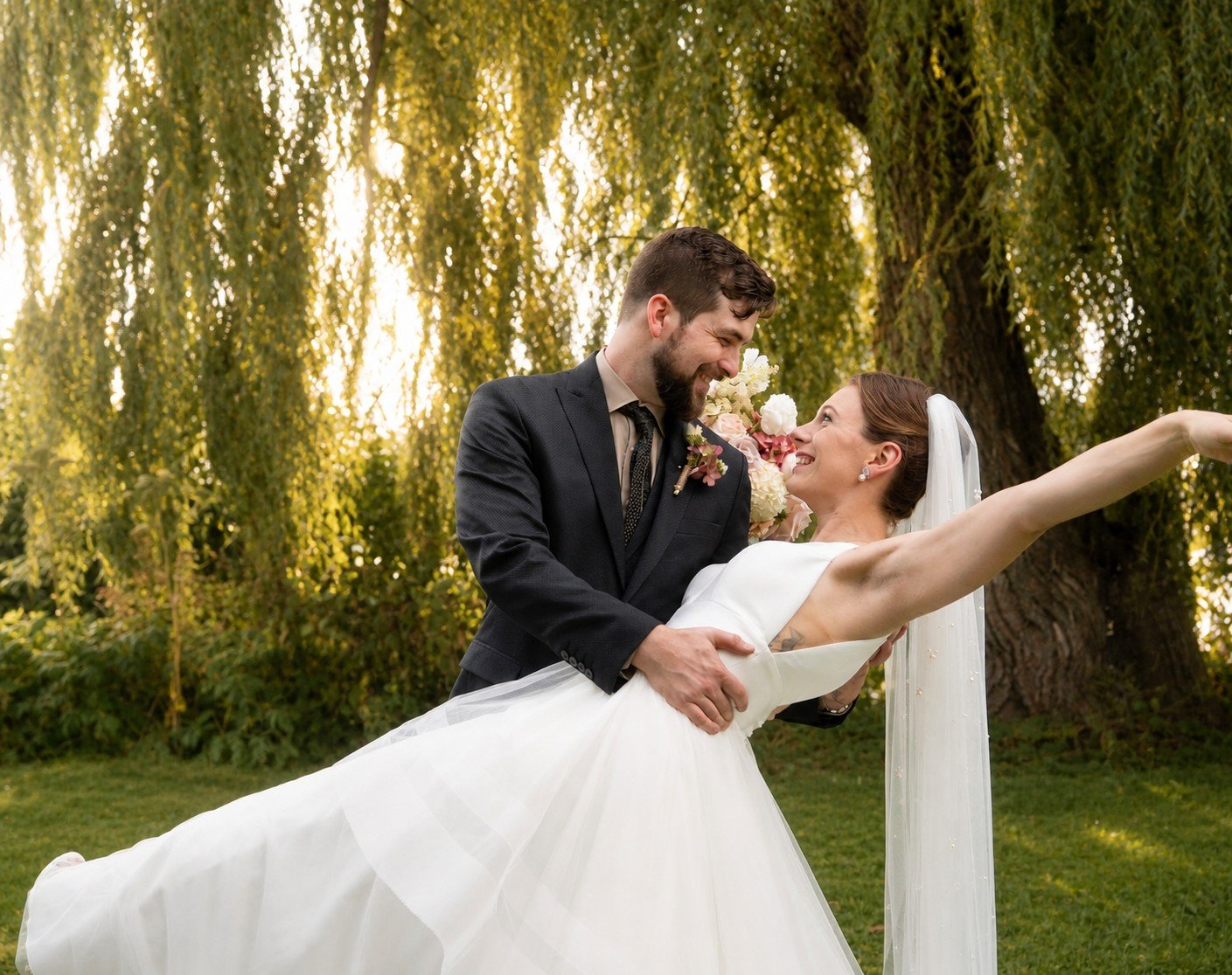 A newlywed couple dancing outdoors with trees in the background, the groom lifting the bride who is in a white wedding dress with a veil and bouquet.