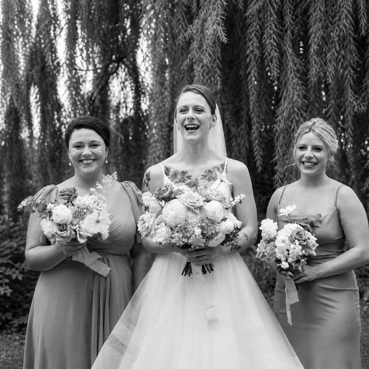 Black and white photo of three women at a wedding, with the bride in the center holding a bouquet, and two bridesmaids, each holding a smaller bouquet, all smiling outdoors in front of trees.