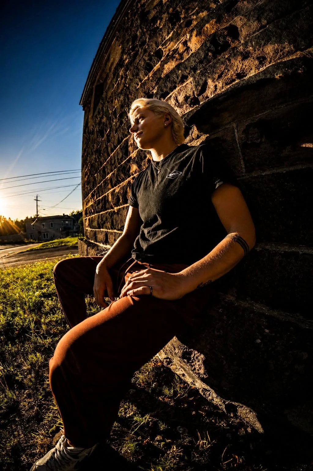 Young woman with blonde hair leaning against a brick wall during sunset, wearing a black t-shirt and brown pants, with a tattooed arm and earrings, outdoors in a suburban area.