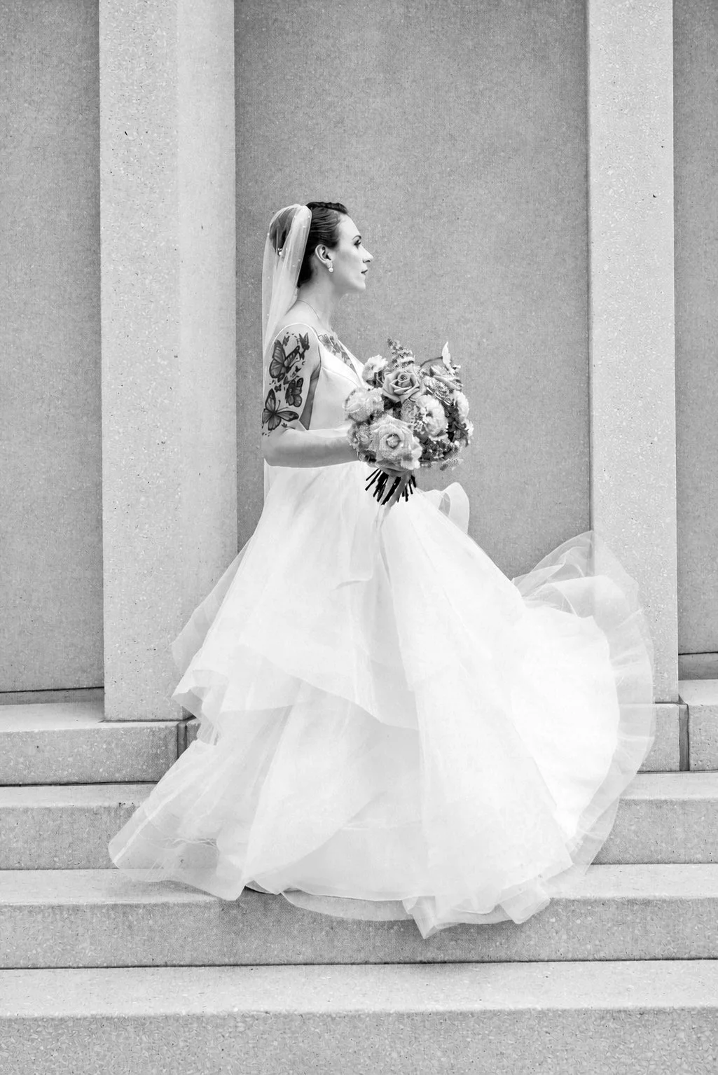 Black and white photo of a bride sitting on steps, holding a bouquet of flowers, wearing a strapless wedding dress with a full tulle skirt, with tattoos on her arm and a veil on her head, against a plain wall background.