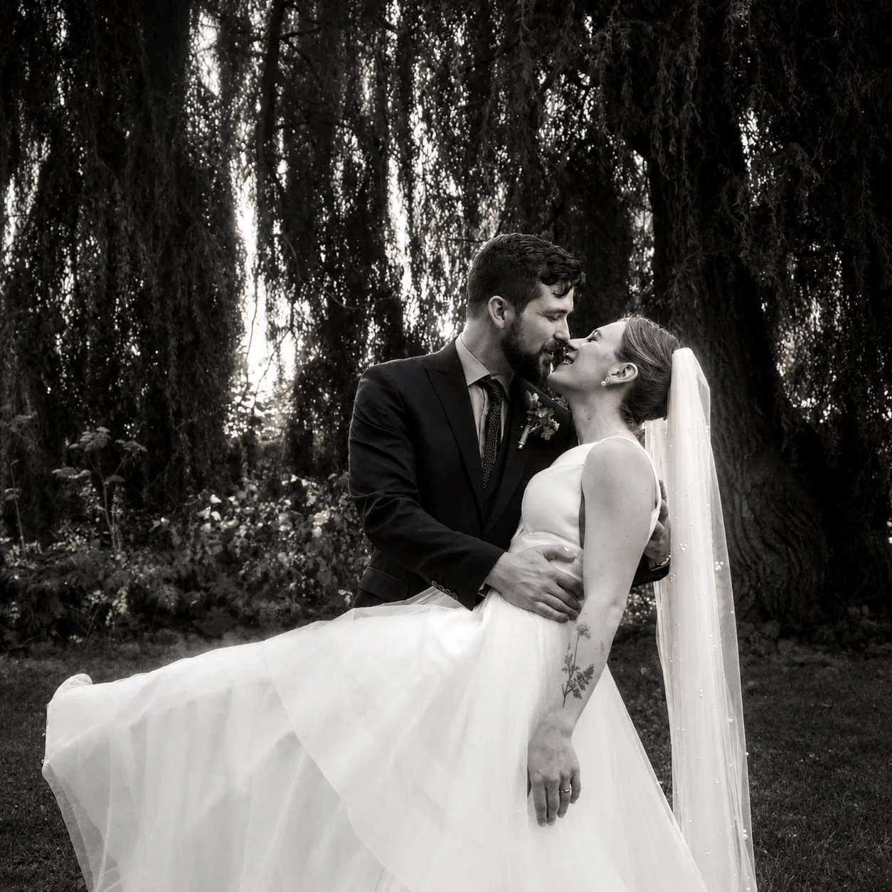 A black and white photo of a bride and groom outdoors, with trees and foliage in the background. The groom is wearing a dark suit and the bride is in a white wedding gown with a veil, and they are sharing a close, romantic moment.
