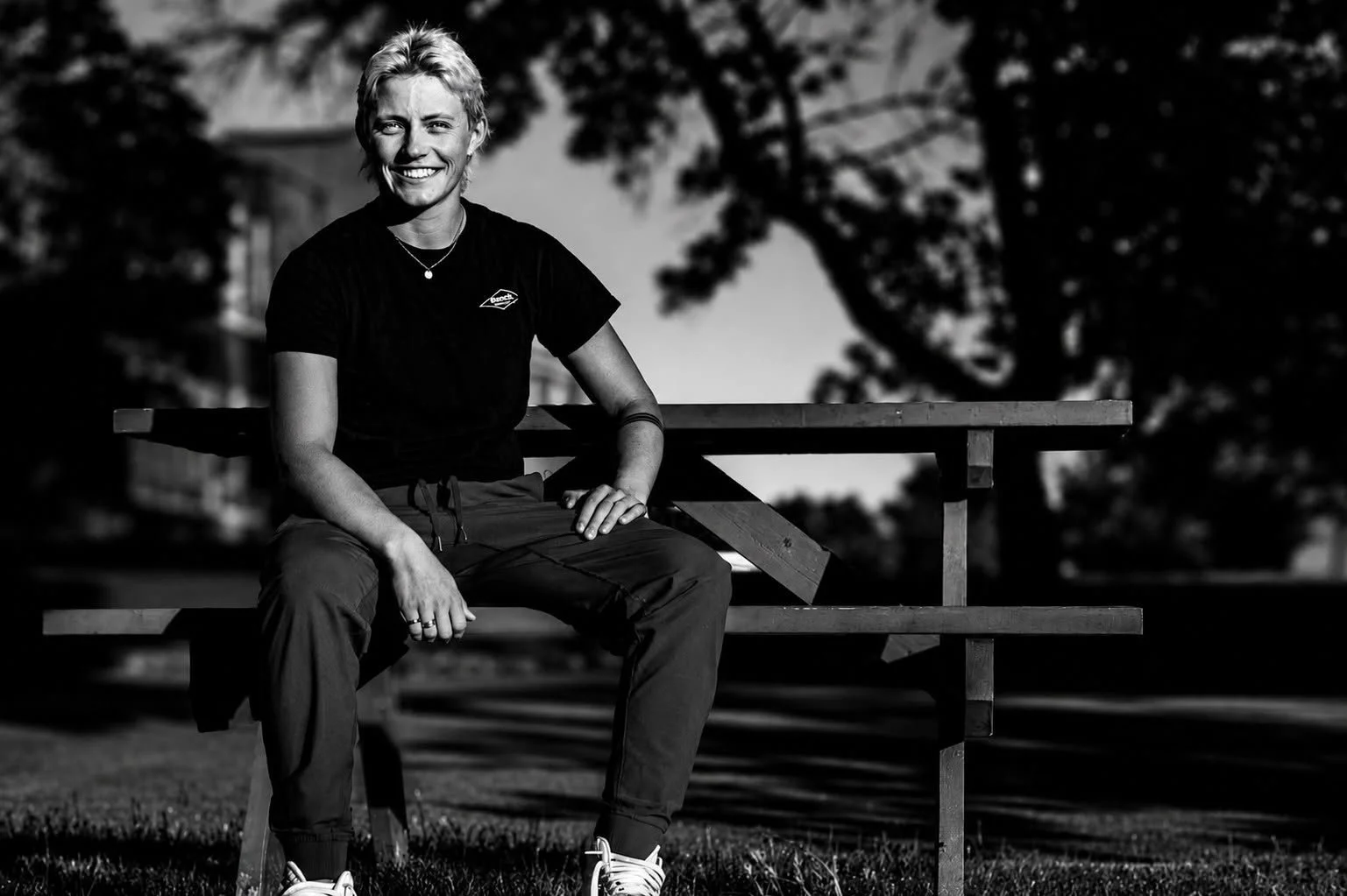 Black and white photo of a smiling woman sitting on a park bench outdoors, with trees in the background.