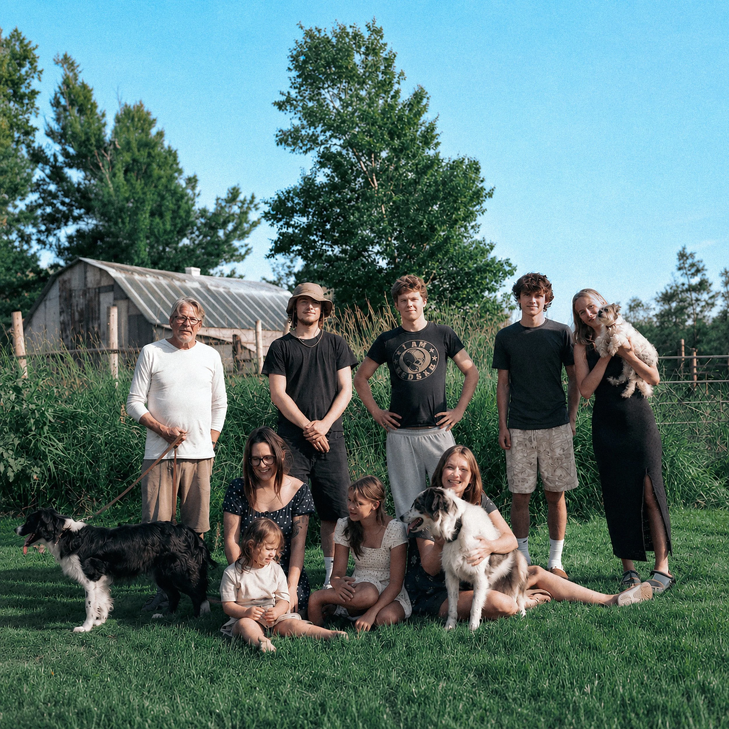 A group of people, including children and adults, outdoors on a grassy area with trees; some holding or with dogs, in a casual setting on a sunny day.