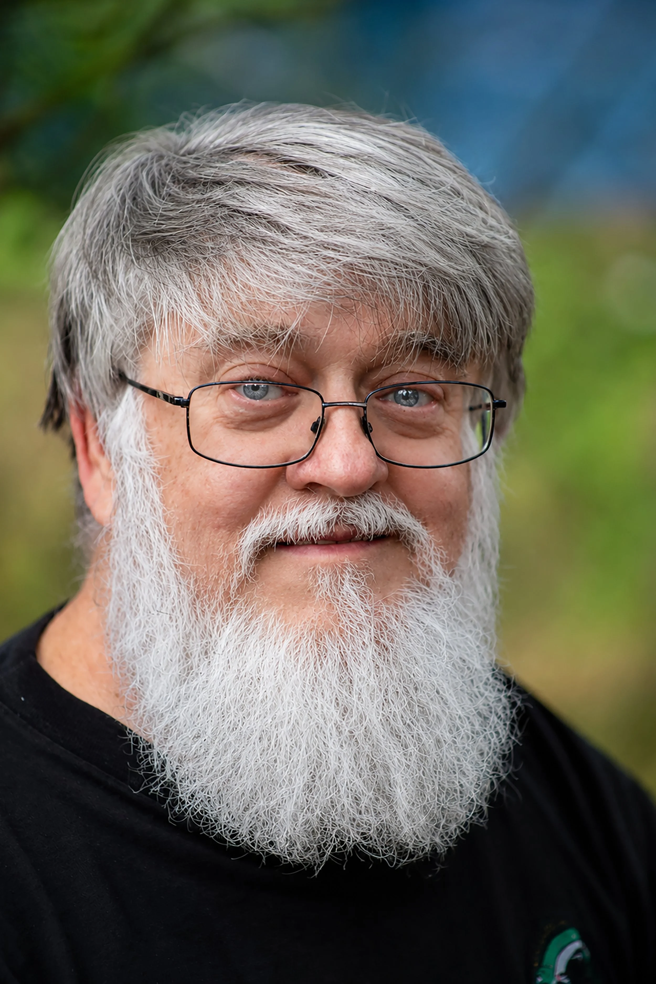 Close-up of an older man with gray hair, glasses, and a long white beard, wearing a black shirt with a small logo, outdoors with a blurred green background.