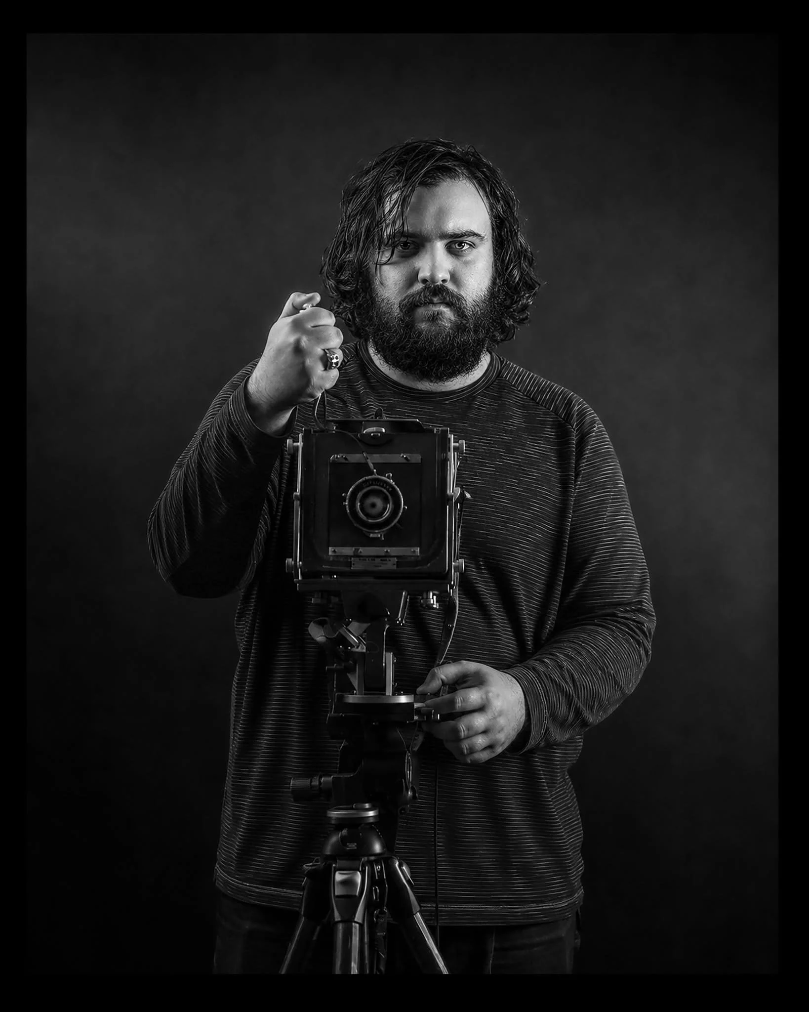 Black and white photo of a man with long hair and beard, wearing a striped shirt, taking a picture with a camera mounted on a tripod, with a focused expression, against a dark background.