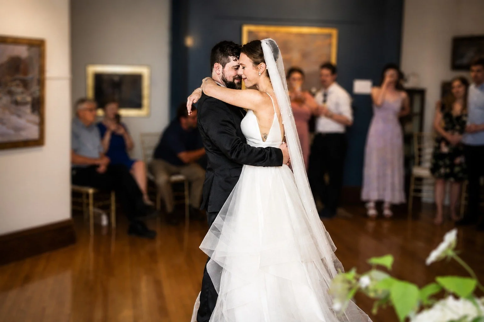 A newlywed couple sharing their first dance at a wedding reception surrounded by seated and standing guests.