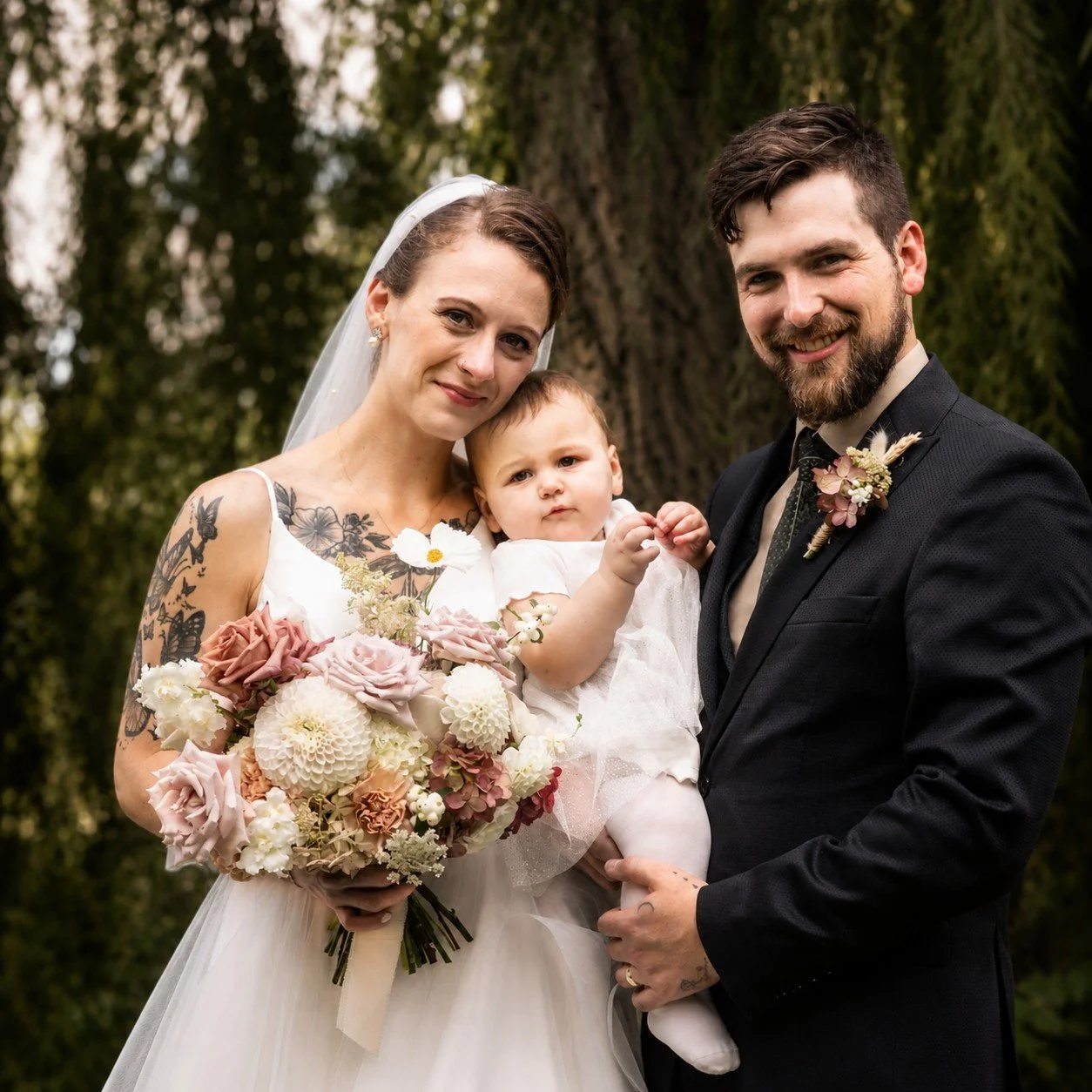 A wedding photo of a bride, groom, and a young child outdoors. The bride wears a wedding dress and veil, holding a large bouquet of flowers. The groom is in a black suit with a boutonniere. The young child, dressed in white, is held by the groom, and