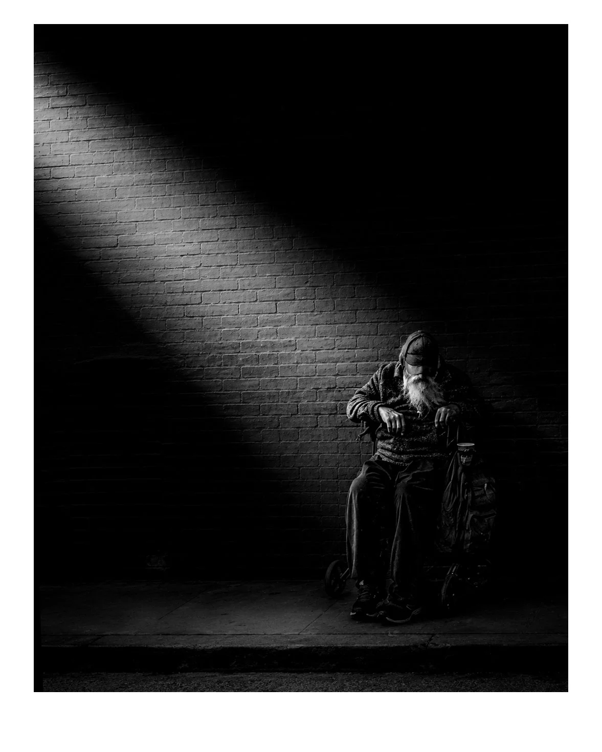 A black and white photo of an elderly man with a beard sitting alone on a chair against a brick wall, illuminated by a single, diagonal spotlight.