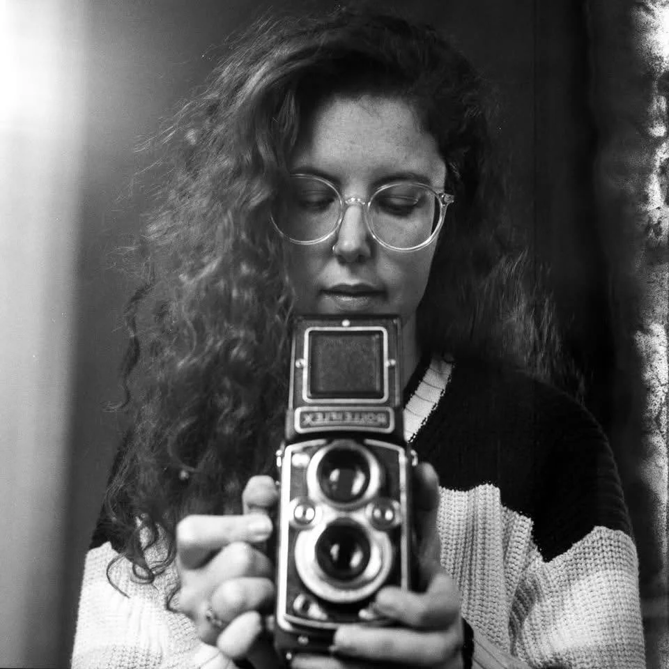 A woman with long curly hair and glasses taking a selfie with a vintage camera in black and white.