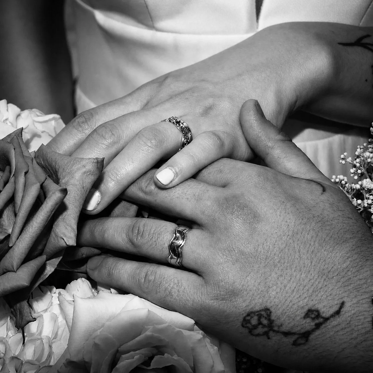 Close-up of two hands with wedding bands resting on each other, surrounded by flowers in black and white.