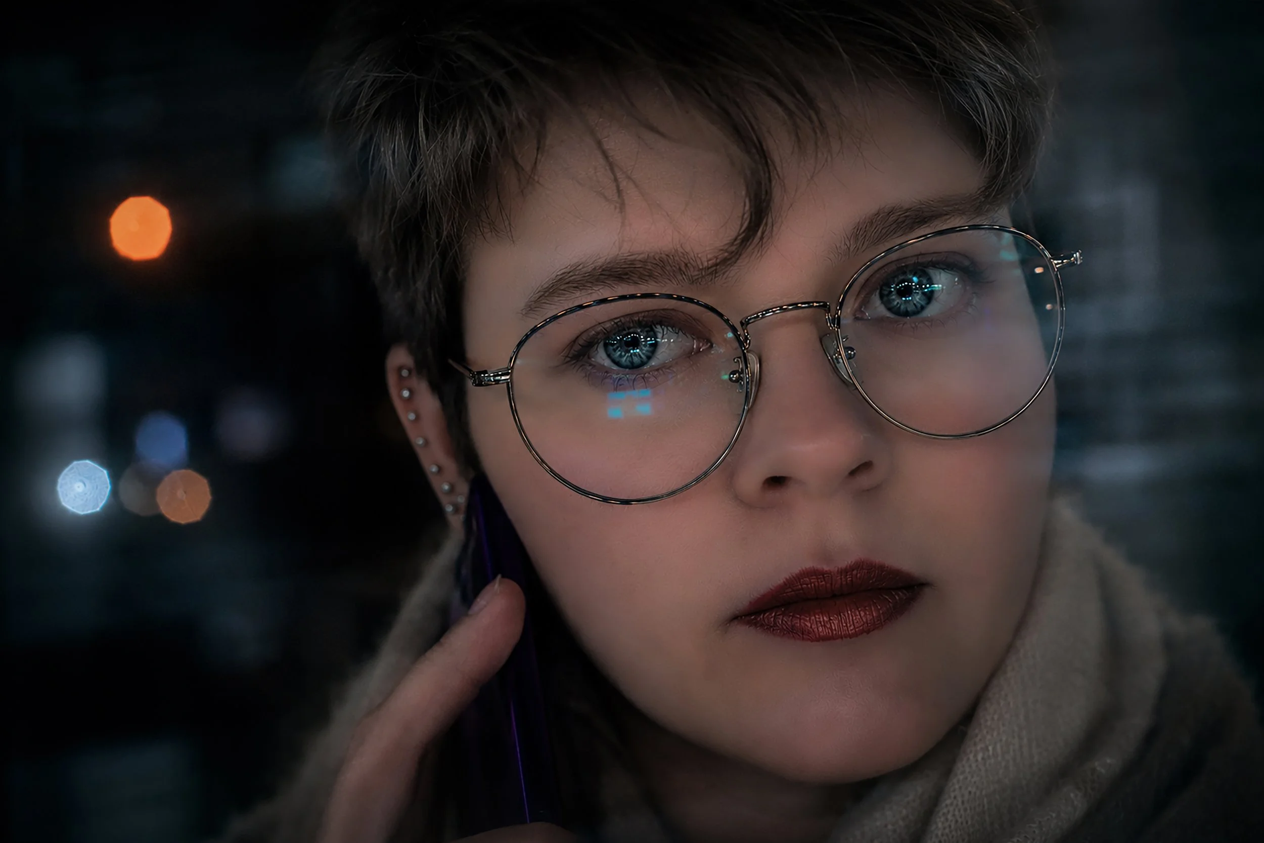 Close-up of a woman with short dark hair and blue eyes wearing round glasses and bold makeup, with city lights blurred in the background.