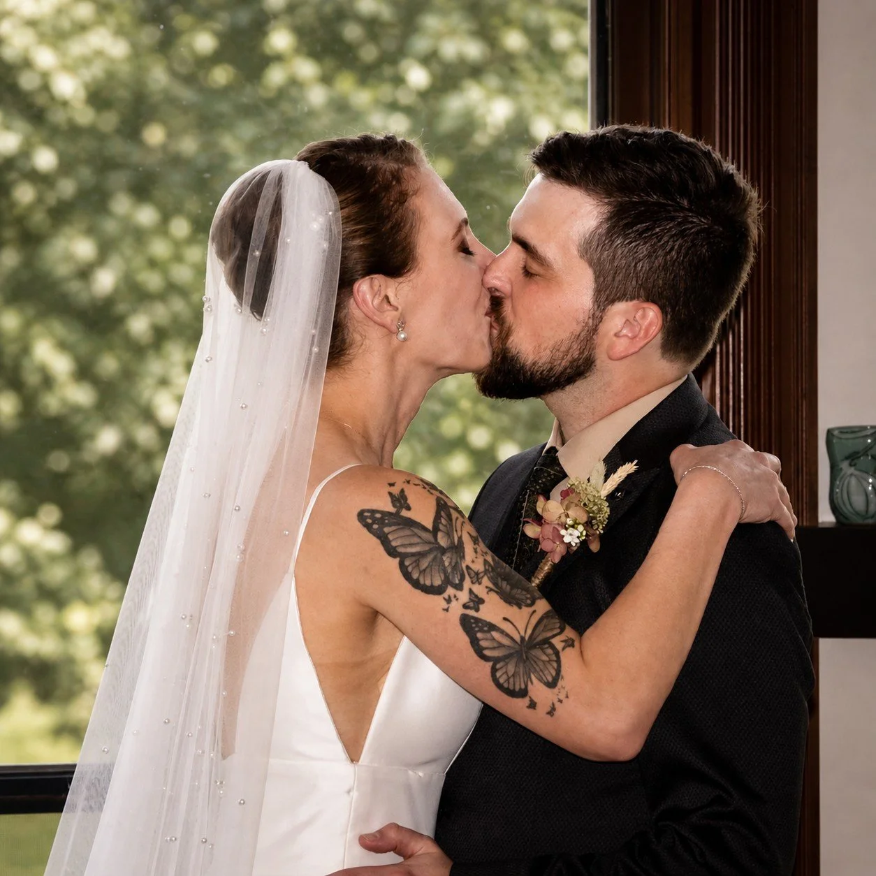 A bride and groom share a kiss during their wedding, with the bride wearing a white dress and veil, and the groom in a black suit, indoors near a window with greenery outside.