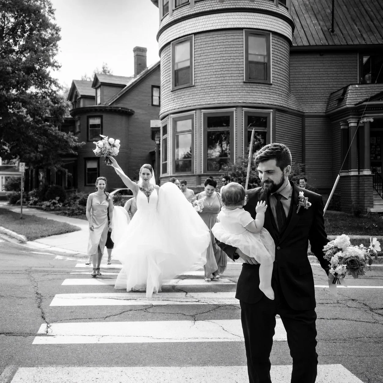 Black and white photo of a wedding procession crossing a street on a crosswalk, with a bride holding a bouquet raising her arm, a man holding a small child, and several women in dresses behind them, all in front of a large Victorian house.