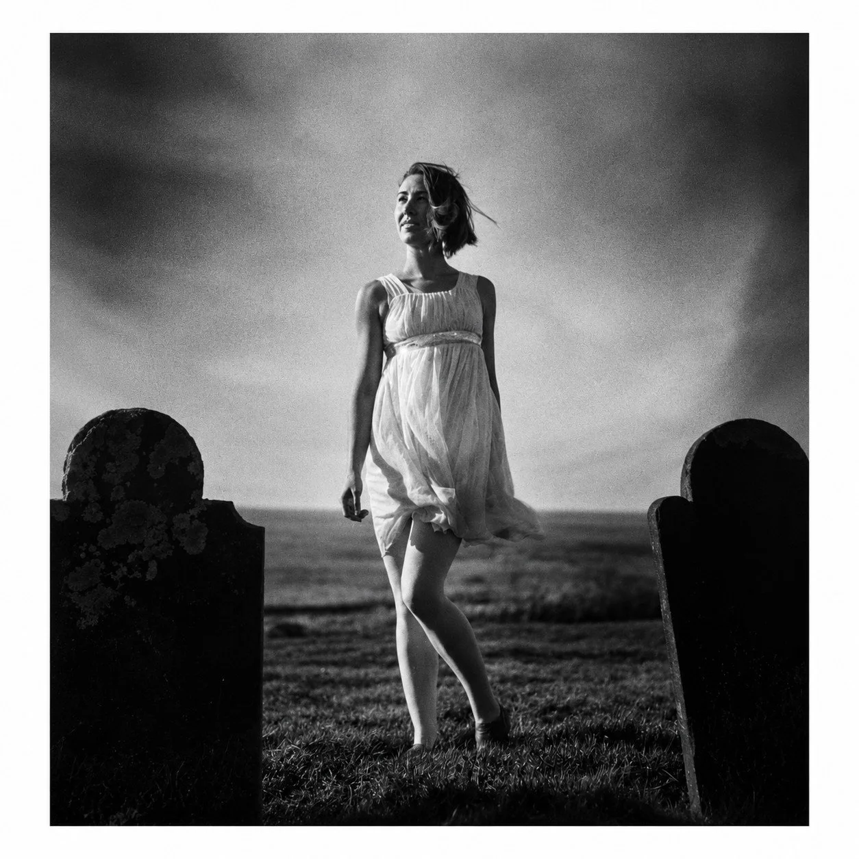 Black and white photo of a woman in a flowing dress standing between two headstones, outdoors near the ocean under a cloudy sky.