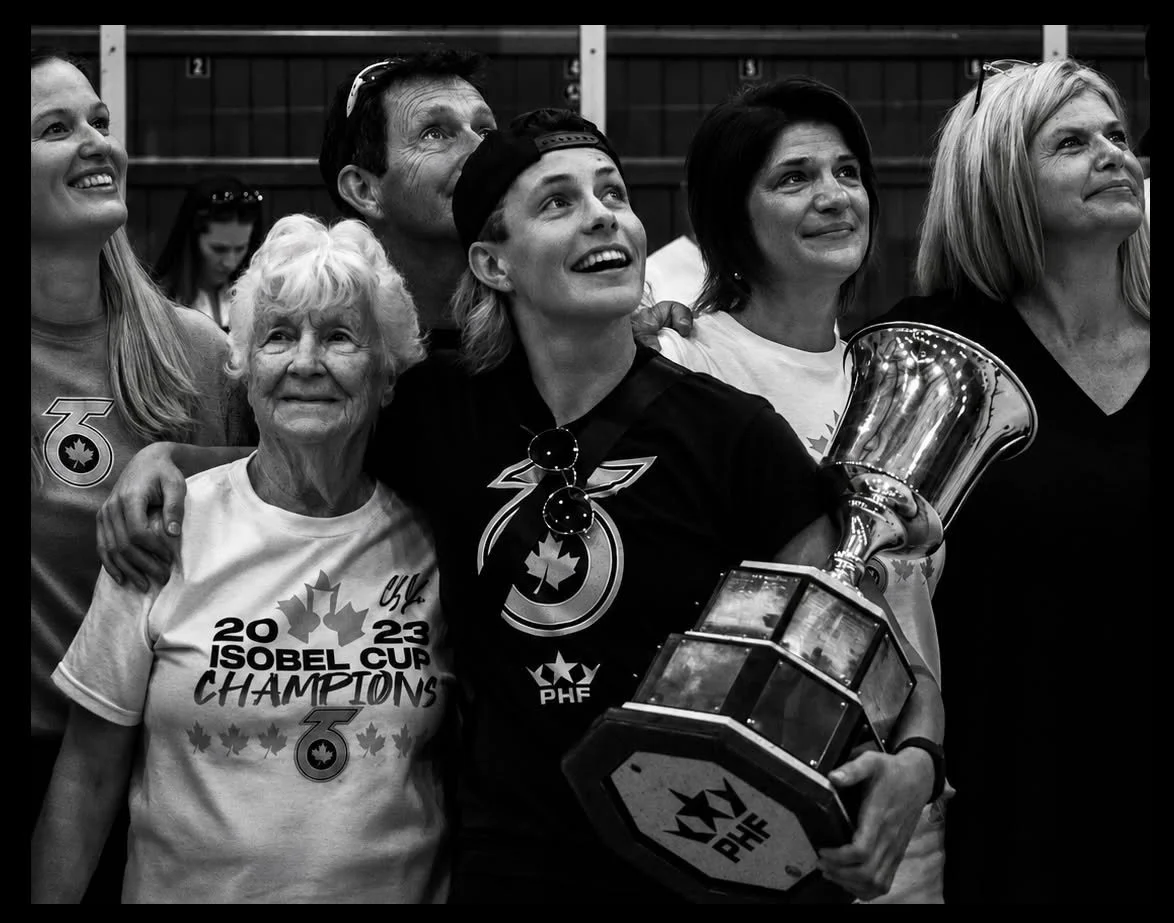 Group of women celebrating with a trophy, including an elderly woman and a young woman wearing a sports jersey, in front of a background with lockers.