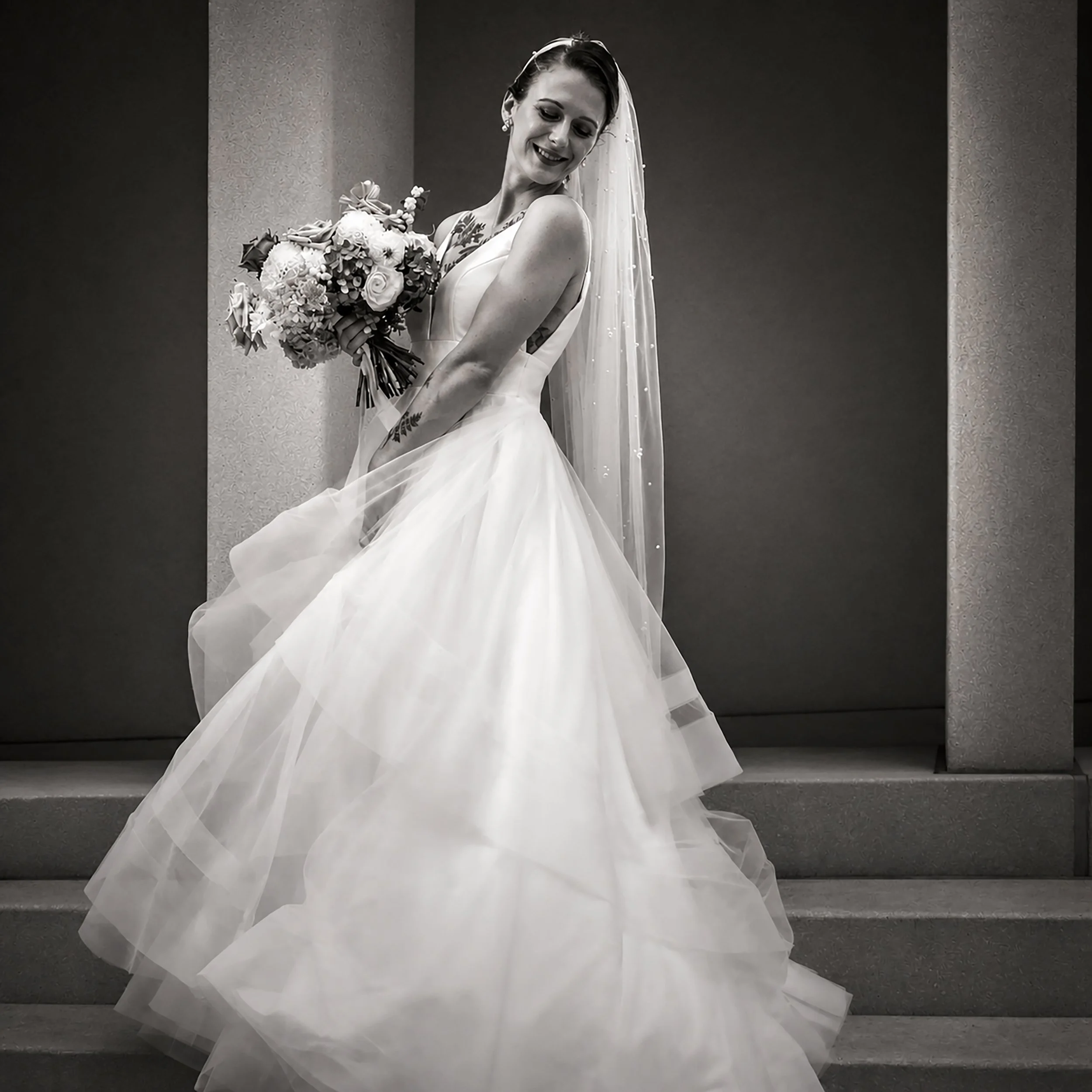Black and white photo of a smiling bride holding a bouquet of flowers, wearing a wedding dress and veil, standing on stairs.