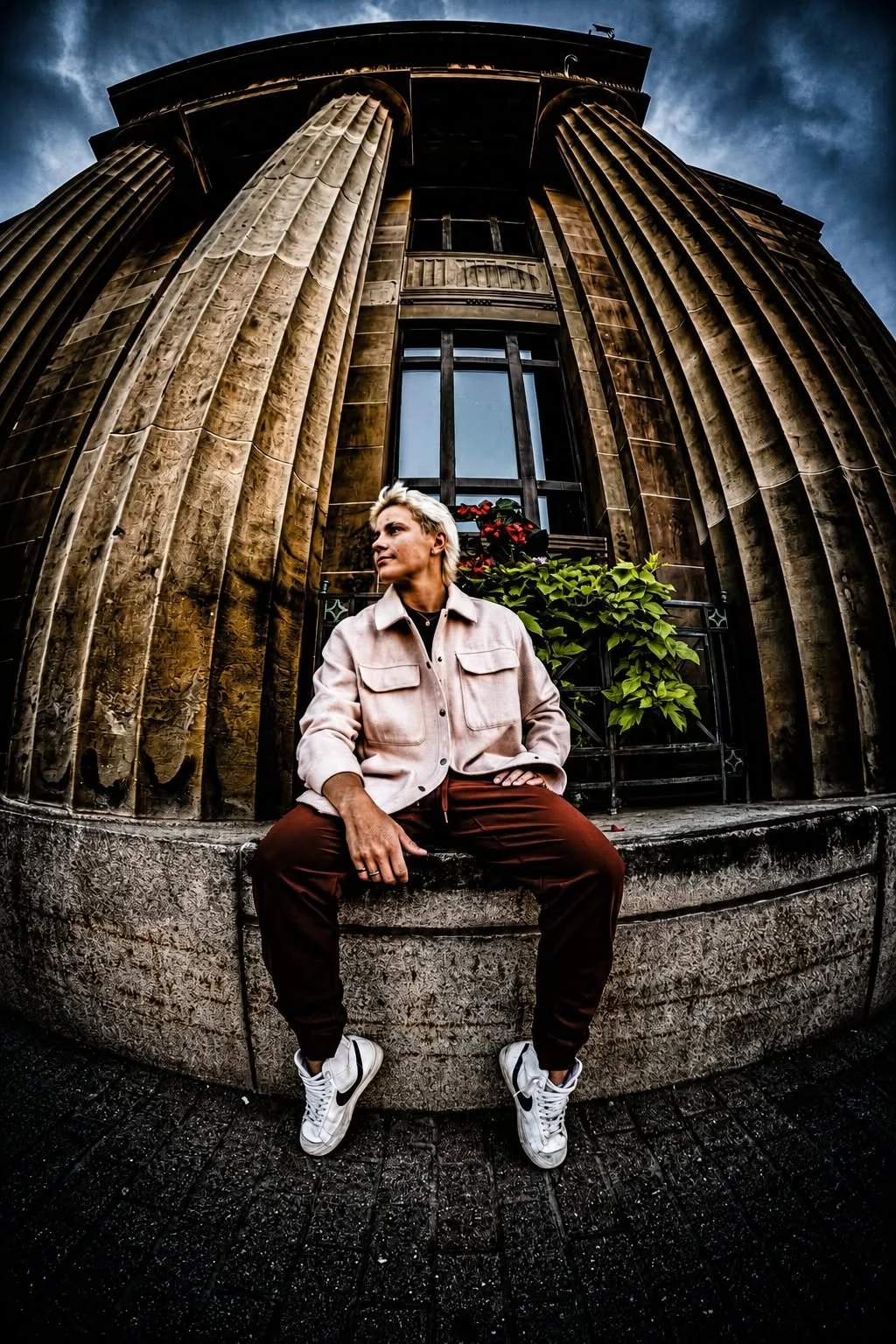 A person with light blond hair sitting on a stone ledge in front of a tall, rounded building with large columns and a dark sky.