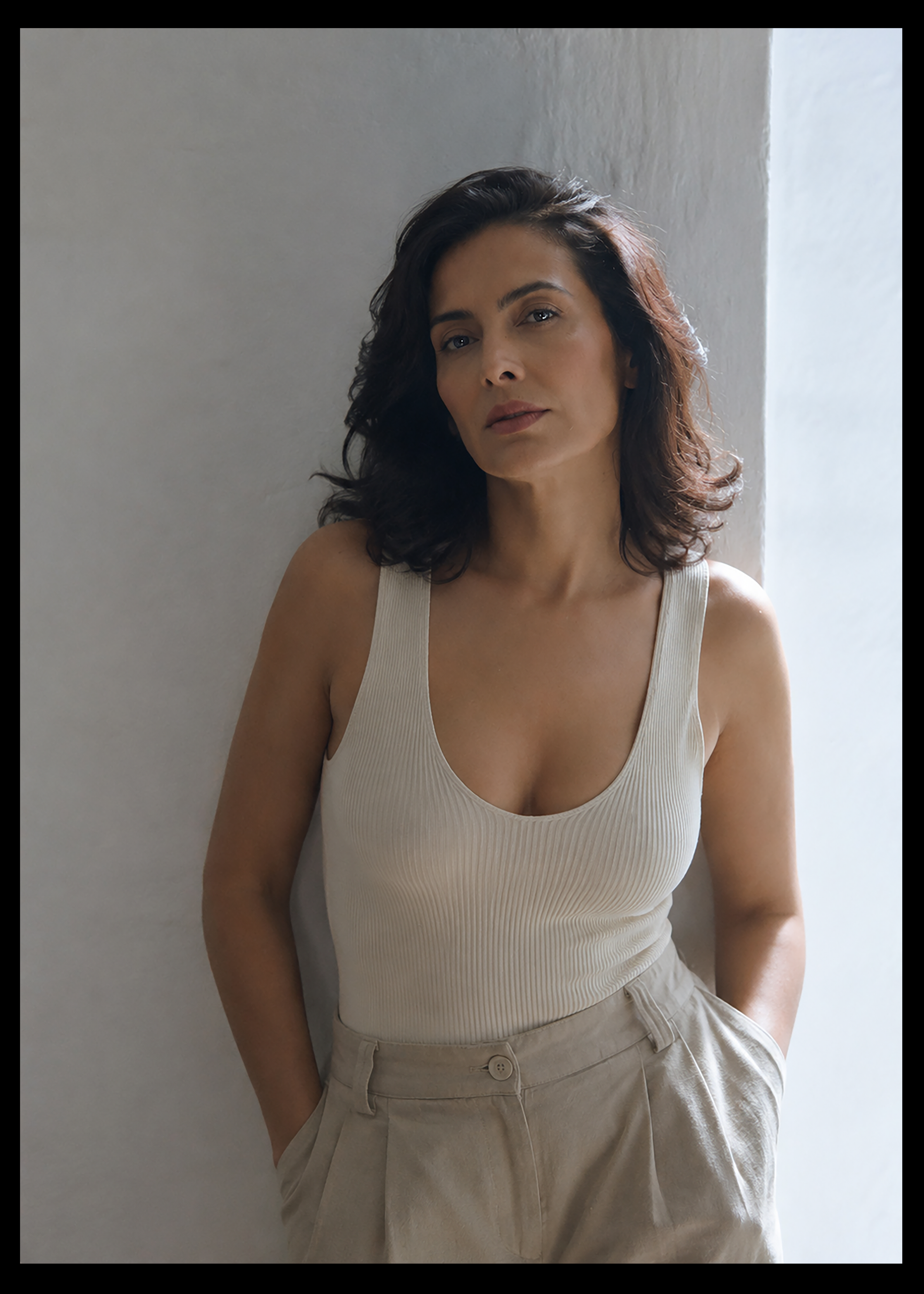 A woman with wavy dark hair wearing a sleeveless beige top and beige pants, standing against a light-colored wall with natural light coming from the right.