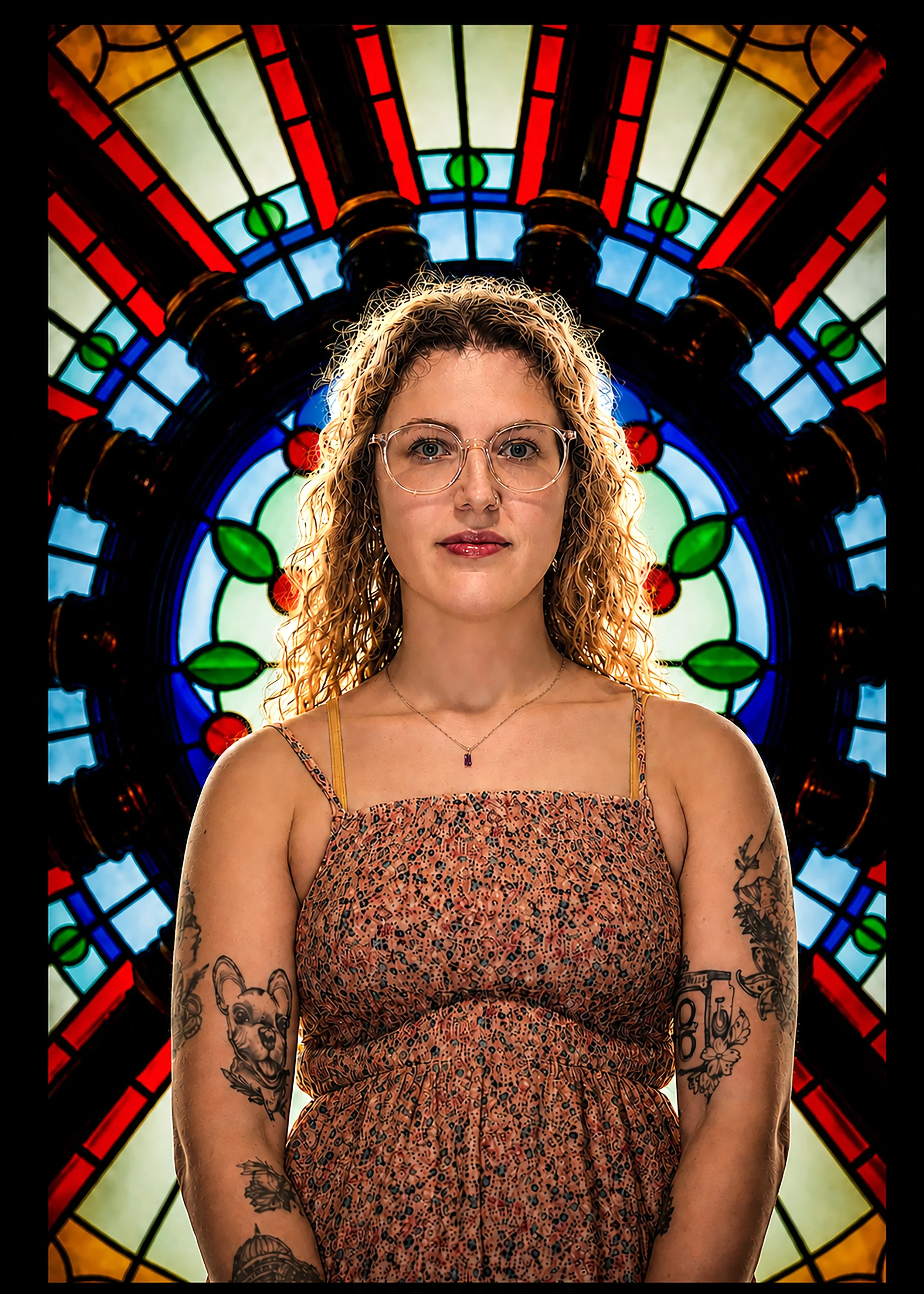 Portrait of a woman with curly hair and glasses, wearing a patterned sleeveless dress, standing in front of a colorful stained glass window.