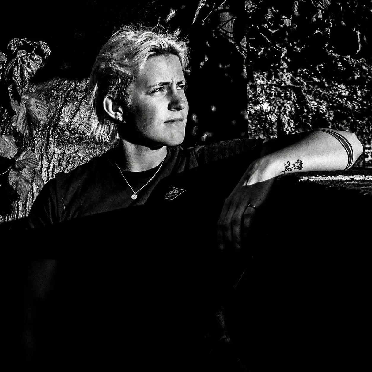 Black and white photo of a woman with short blonde hair, sitting outdoors next to a tree, gazing thoughtfully into the distance with her arm resting on a surface.