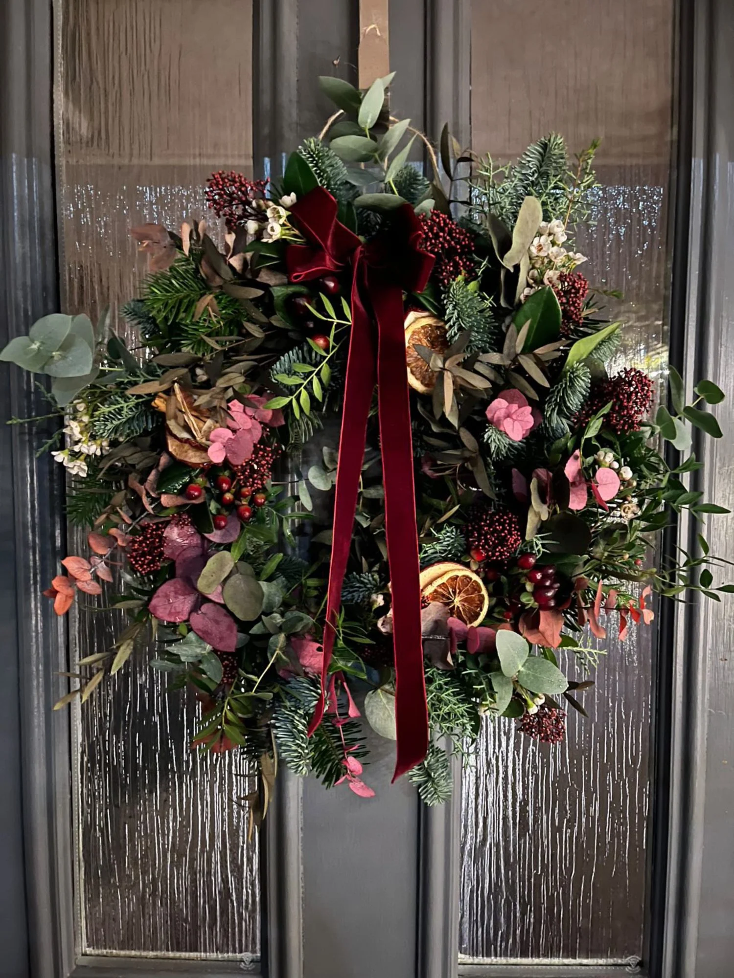 Natural handmade Christmas door wreath with seasonal greenery, dried orange slices and burgundy velvet ribbon displaying on a front door