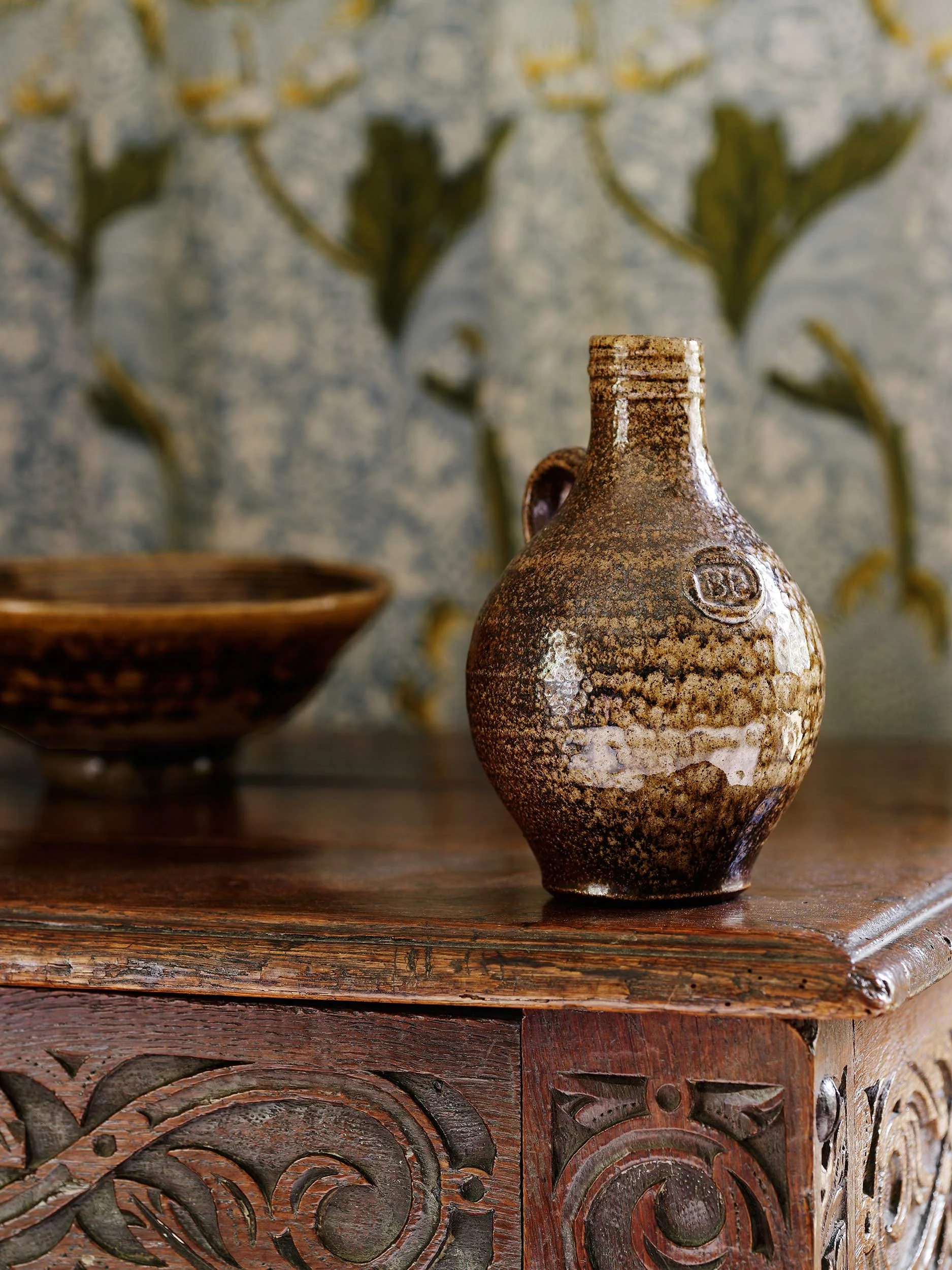 A rustic ceramic vase with a textured brown glaze sits on a wooden surface, with a blurred decorative background featuring leaf and vine patterns.