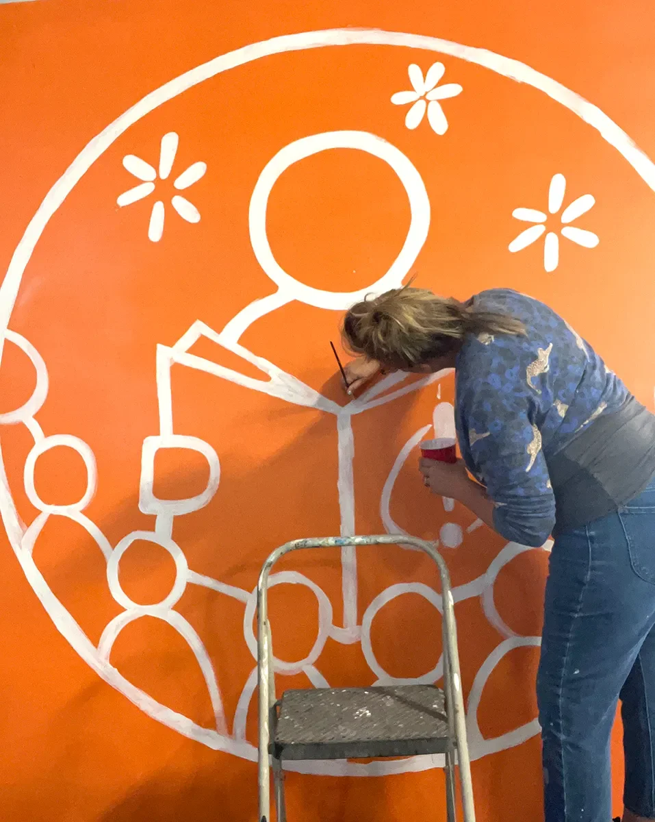 Person painting a white outline of a geometric and floral design on an orange wall, using a small paintbrush and holding a bottle of white paint, standing on a black step stool.