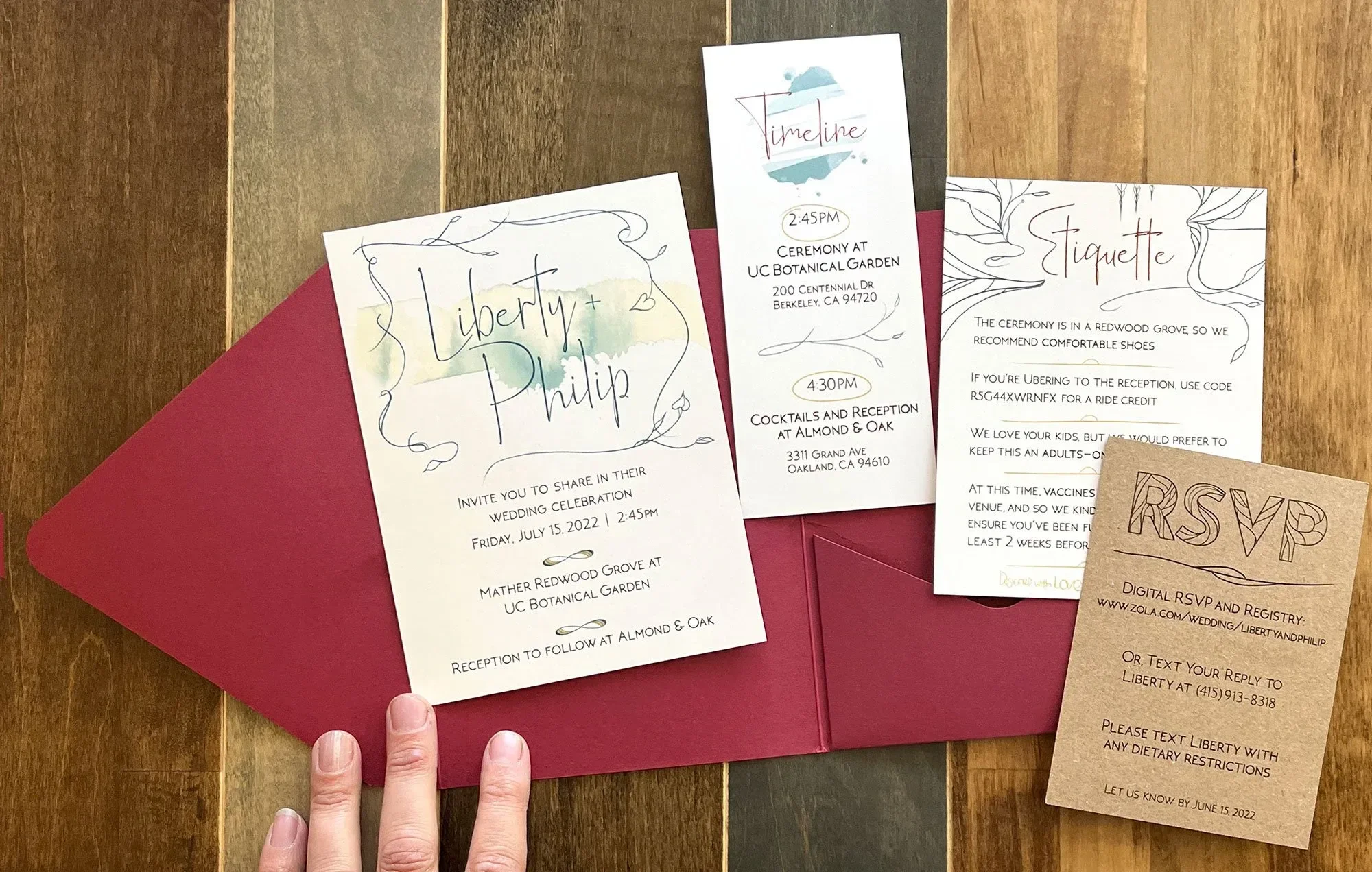 Wedding invitation set including an invitation for Liberty and Philip's wedding, a timeline card, a etiquette card, and an RSVP card, all placed on a wooden surface with a red envelope.
