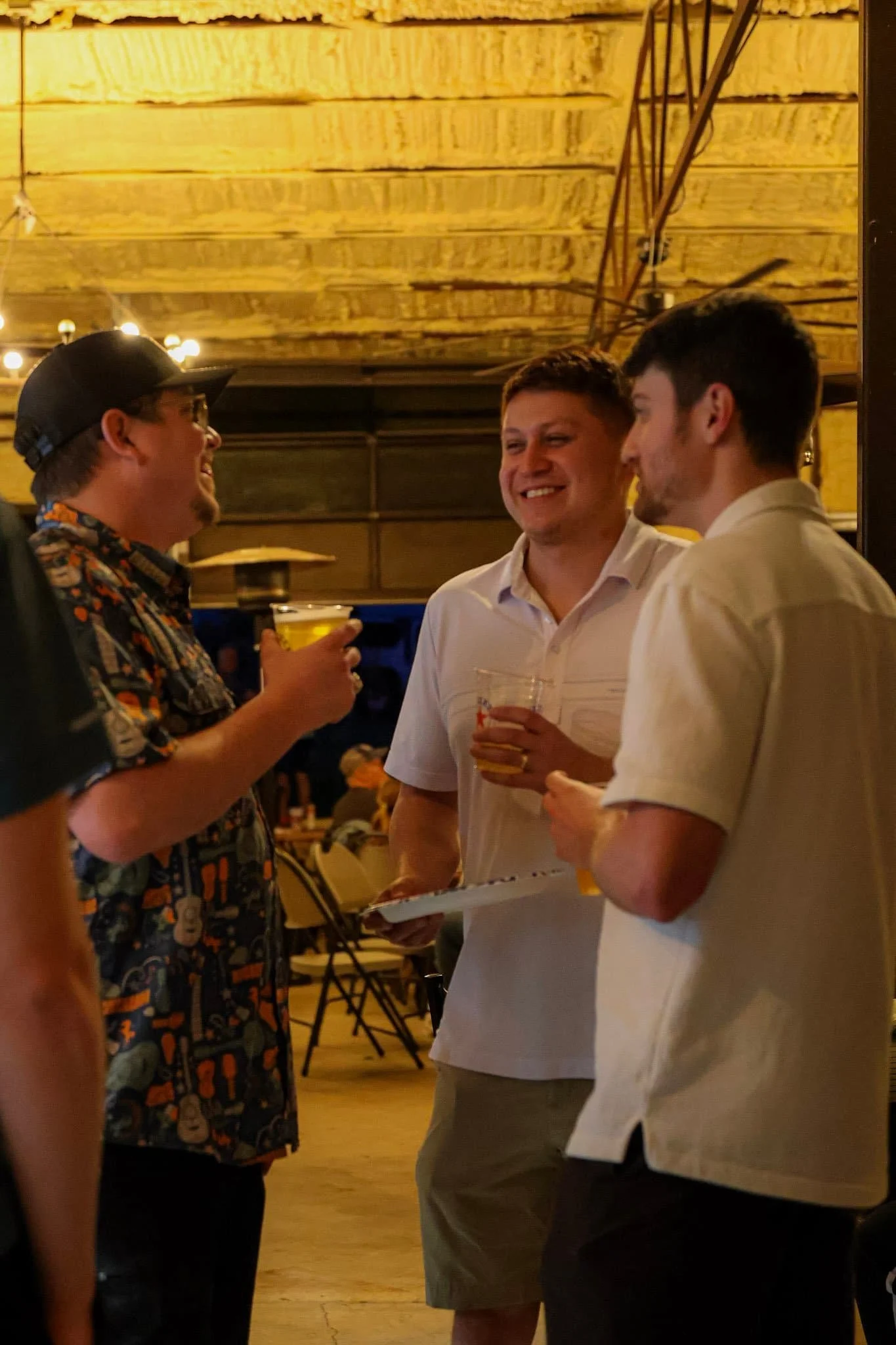 Three men stand together indoors, smiling and holding drinks, chatting about the upcoming Summer Concert at a casual gathering.