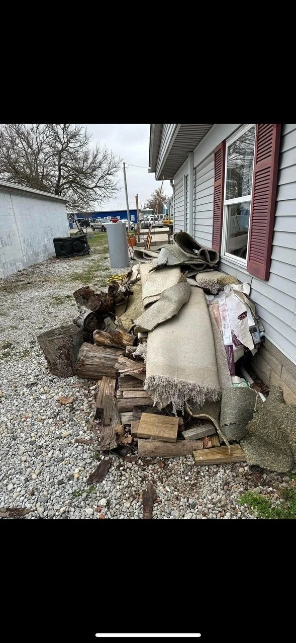 Pile of debris including broken wood, carpet, and other materials outside a house near a window with red shutters.