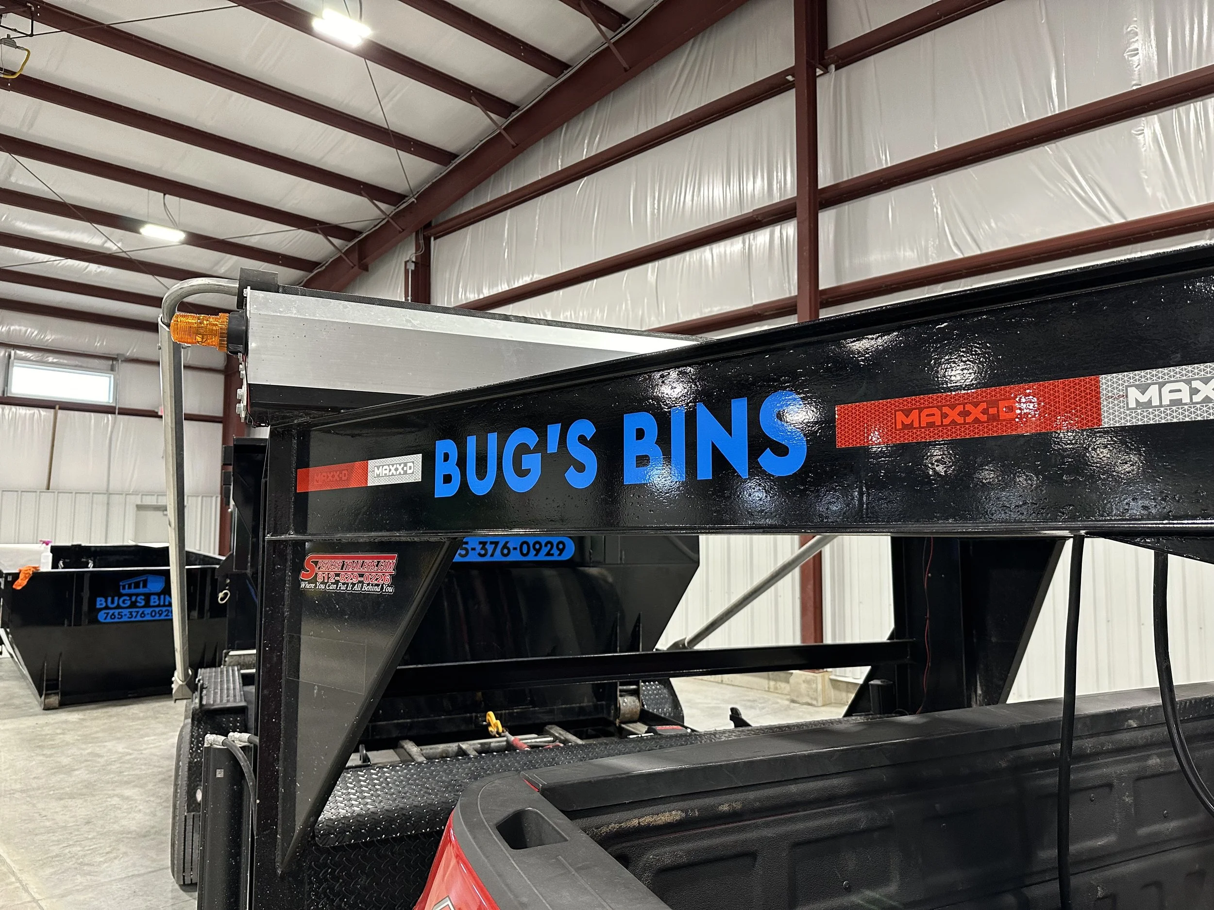 Black utility trailer with blue lettering that reads 'BUG'S BINS' parked inside a warehouse with metal walls and ceiling.
