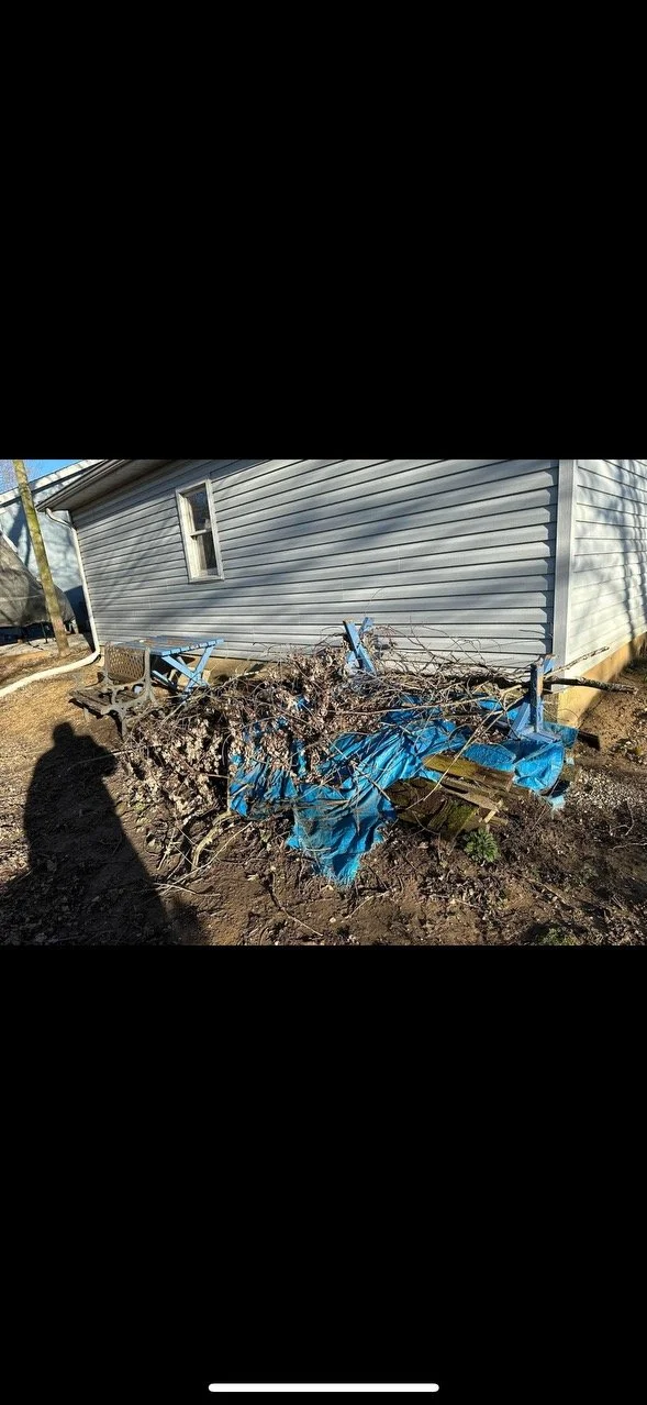 Side view of a house with vinyl siding and a small window, and a pile of debris including branches, blue tarp, and pieces of wood on the ground