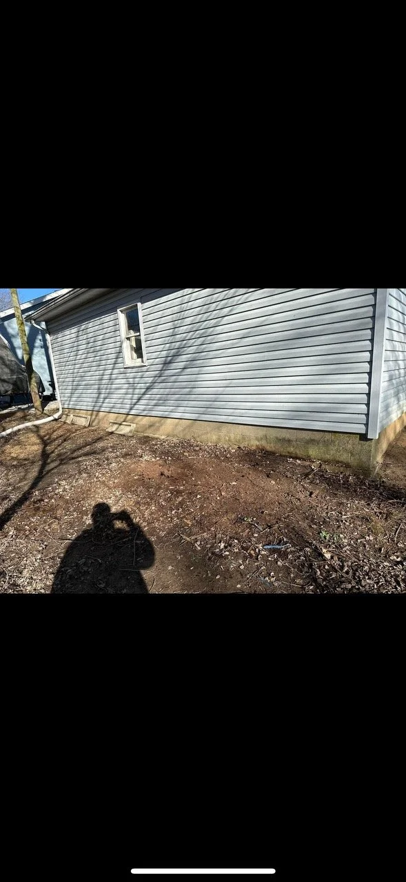 Side of a house with gray vinyl siding, a small window, and a dirt yard with some debris.