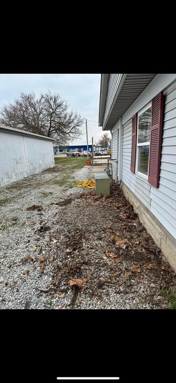 Side yard of a house with gravel ground, fallen leaves, and a gray utility box. The house has white siding with burgundy shutters, and a window. In the background, there are parked cars, a tree, and a clear sky.