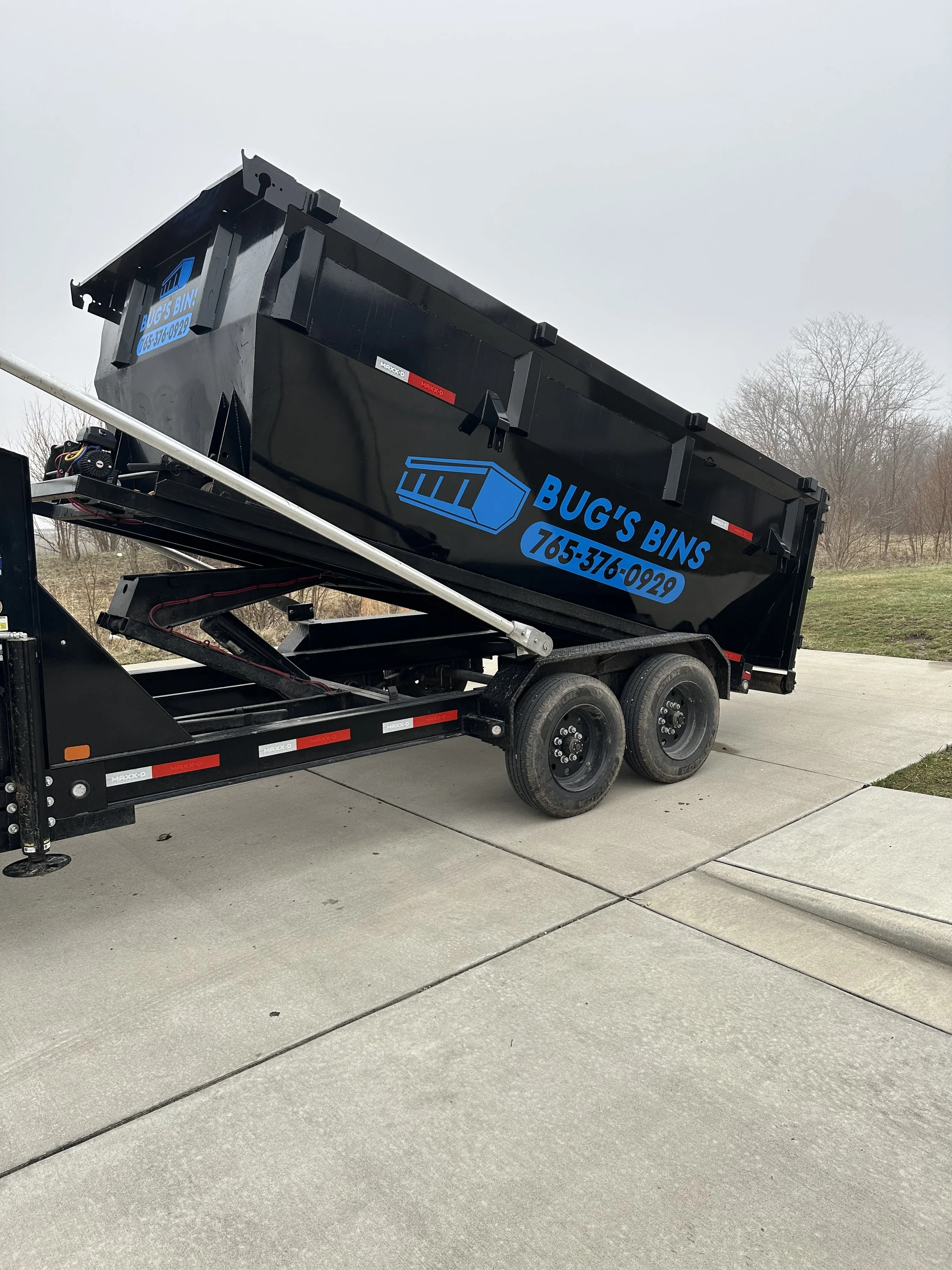 Black dump trailer with blue logo and text reading 'Bug's Bins 765-376-0929' parked on driveway, trailer tilted for dumping.
