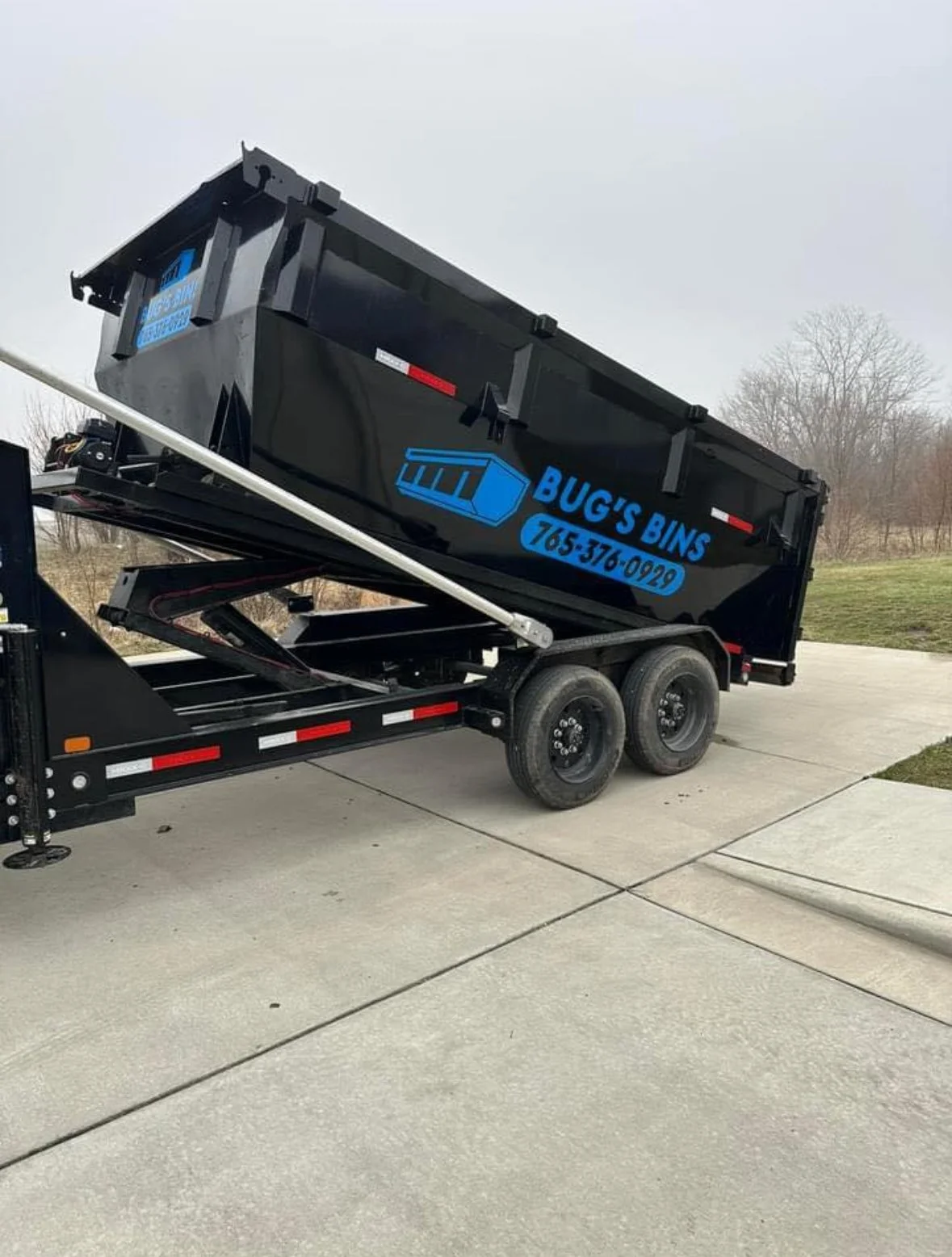 A black dumpster trailer with the text "Bug's Bins" and a phone number, partially raised on a trailer in a parking lot.
