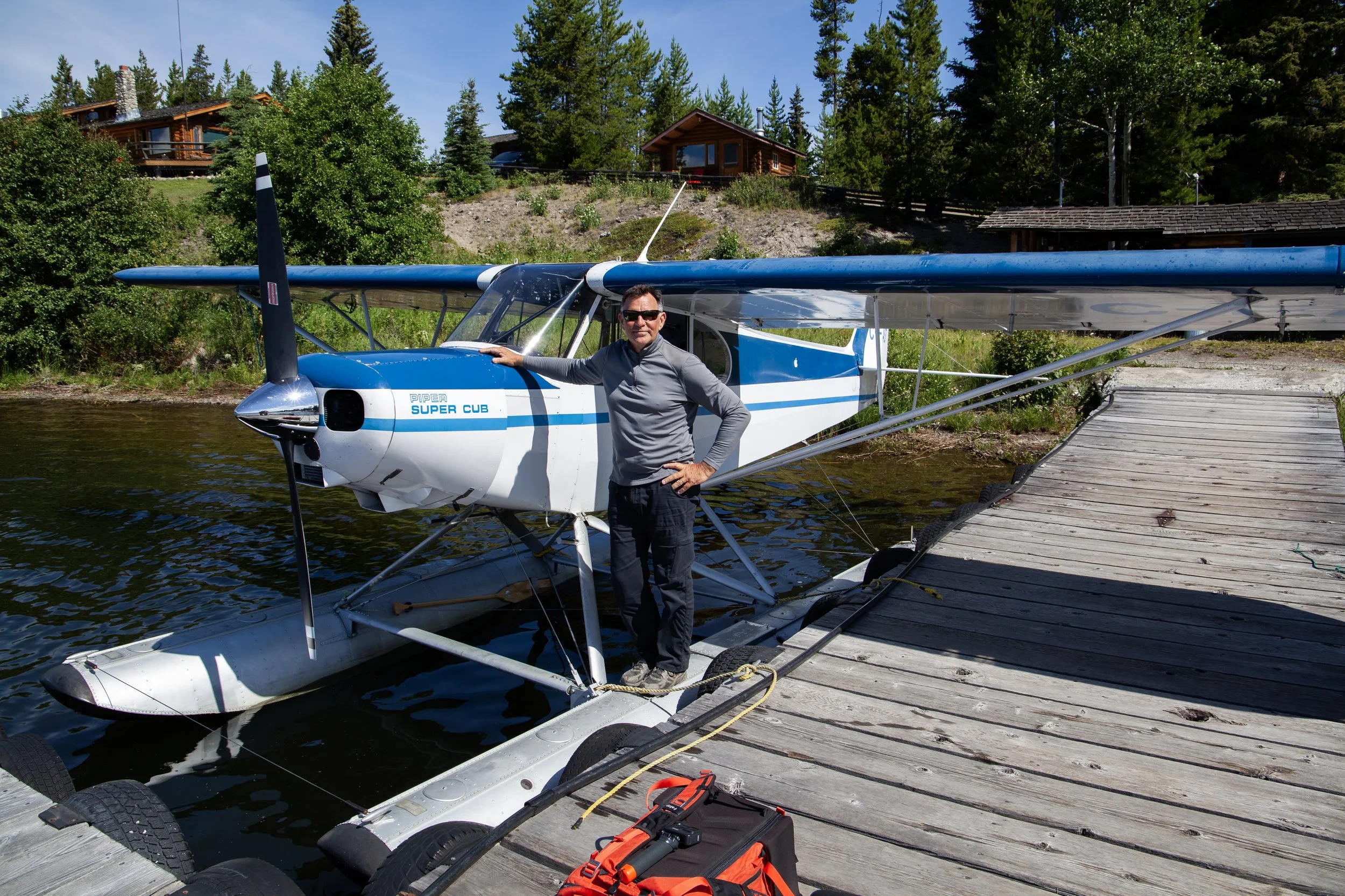 A man standing on a dock next to a seaplane by a lake, with houses and trees in the background.