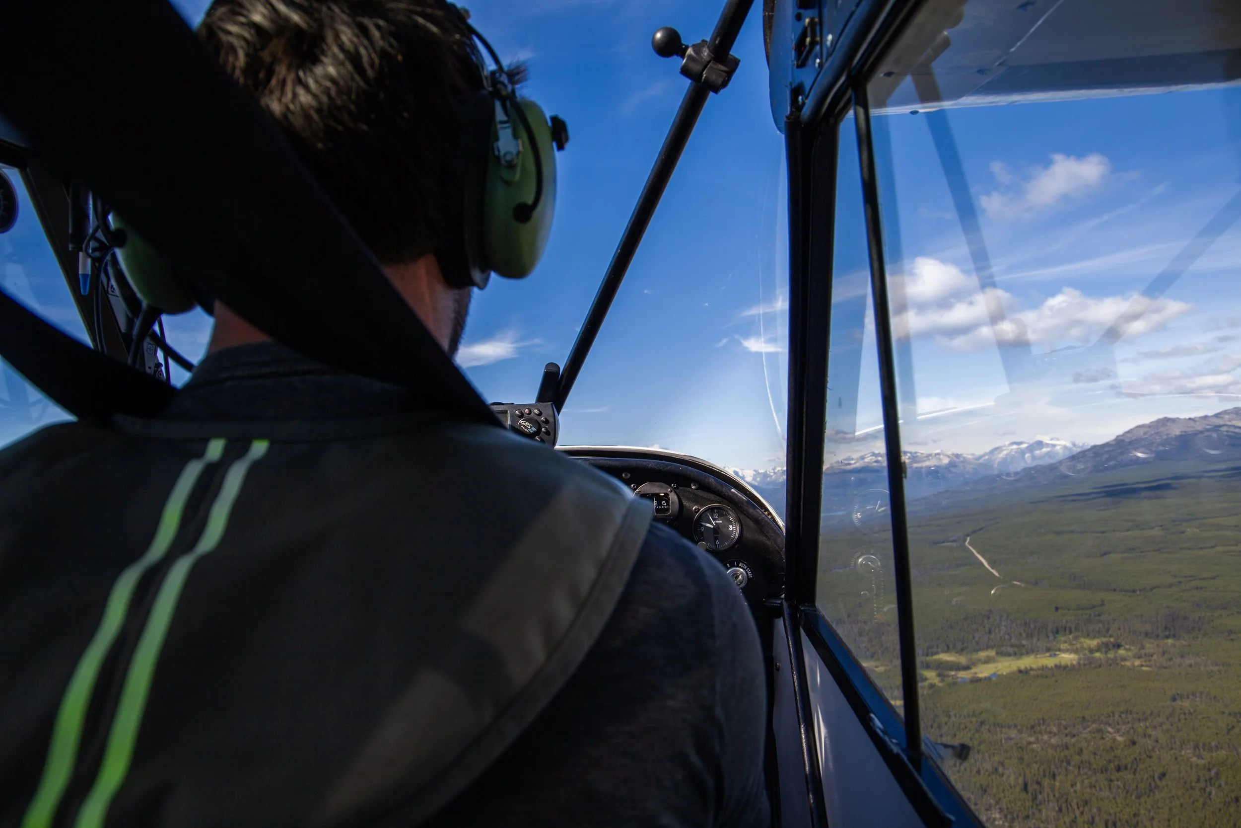 A pilot flying a small airplane over a green landscape with mountains in the distance, wearing a headset and looking forward through the cockpit window.