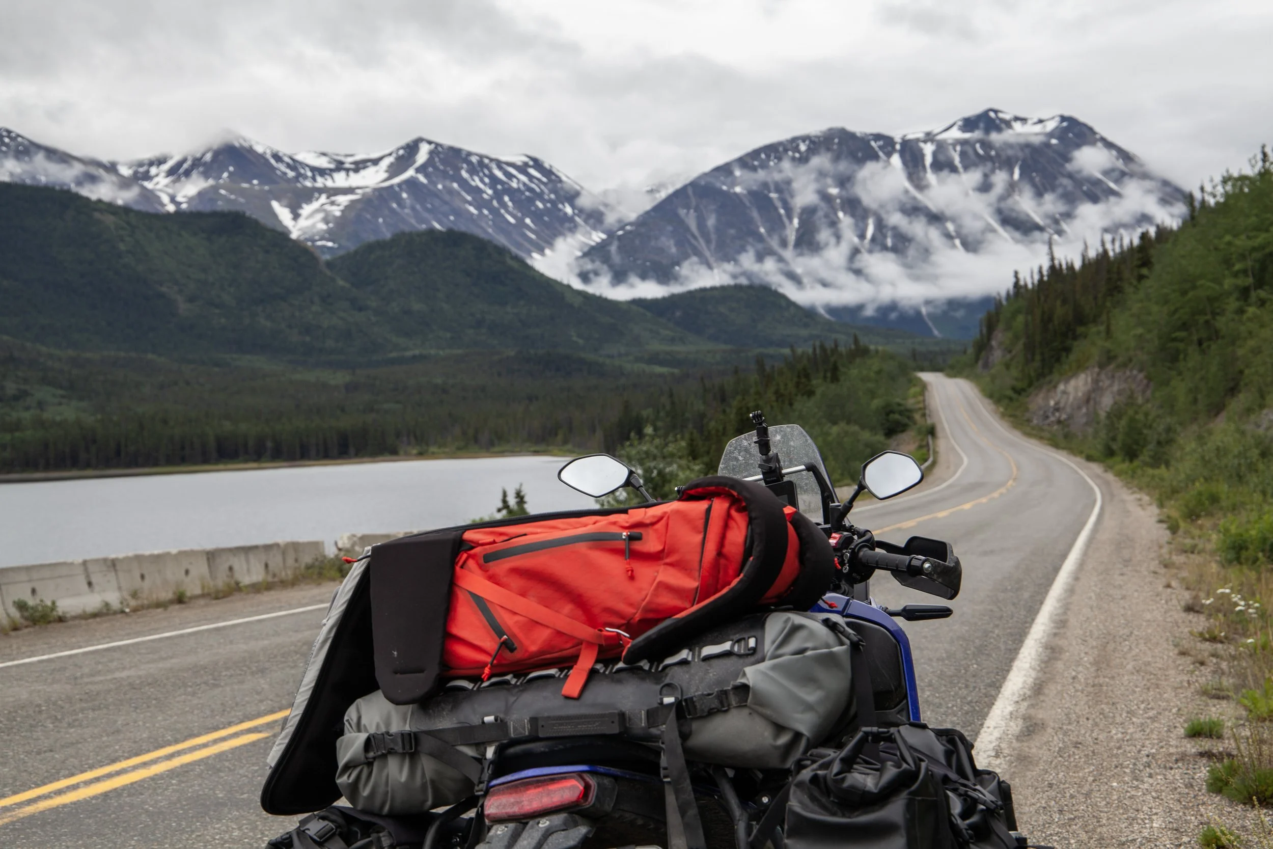 Motorcycle with red and gray backpacks parked on a scenic mountain road with snow-capped peaks, green hills, and a lake in the background.
