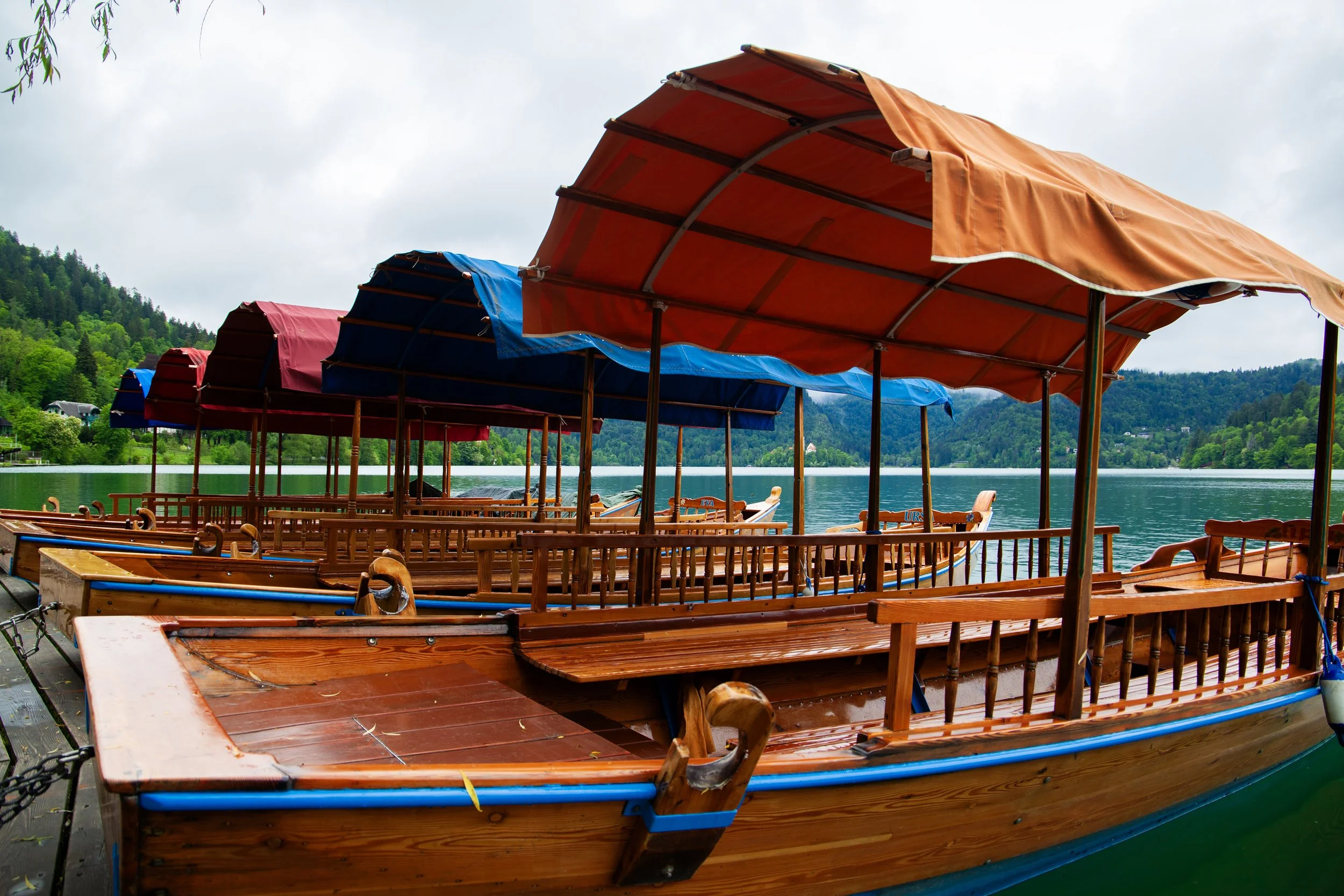 Multiple wooden boats with colorful canopies docked by a lakeside with green trees and hills in the background.