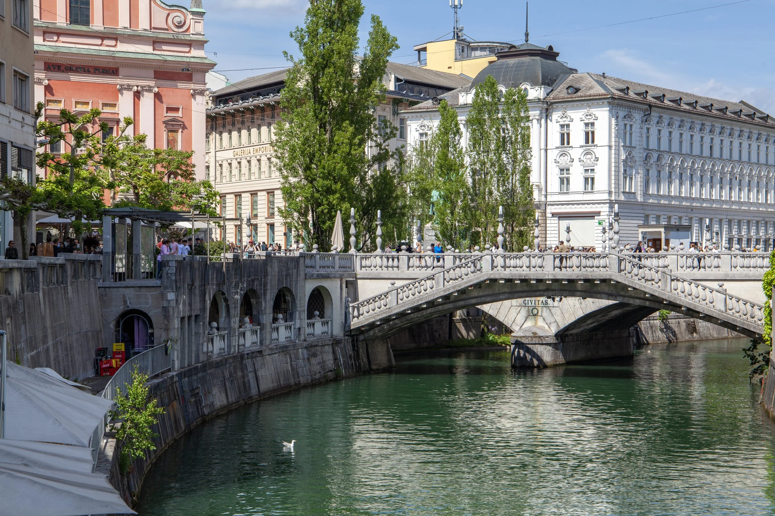 View of a city canal with a stone bridge, surrounded by historic buildings and green trees. People are walking along the bridge and riverside, with some umbrellas and outdoor seating visible.