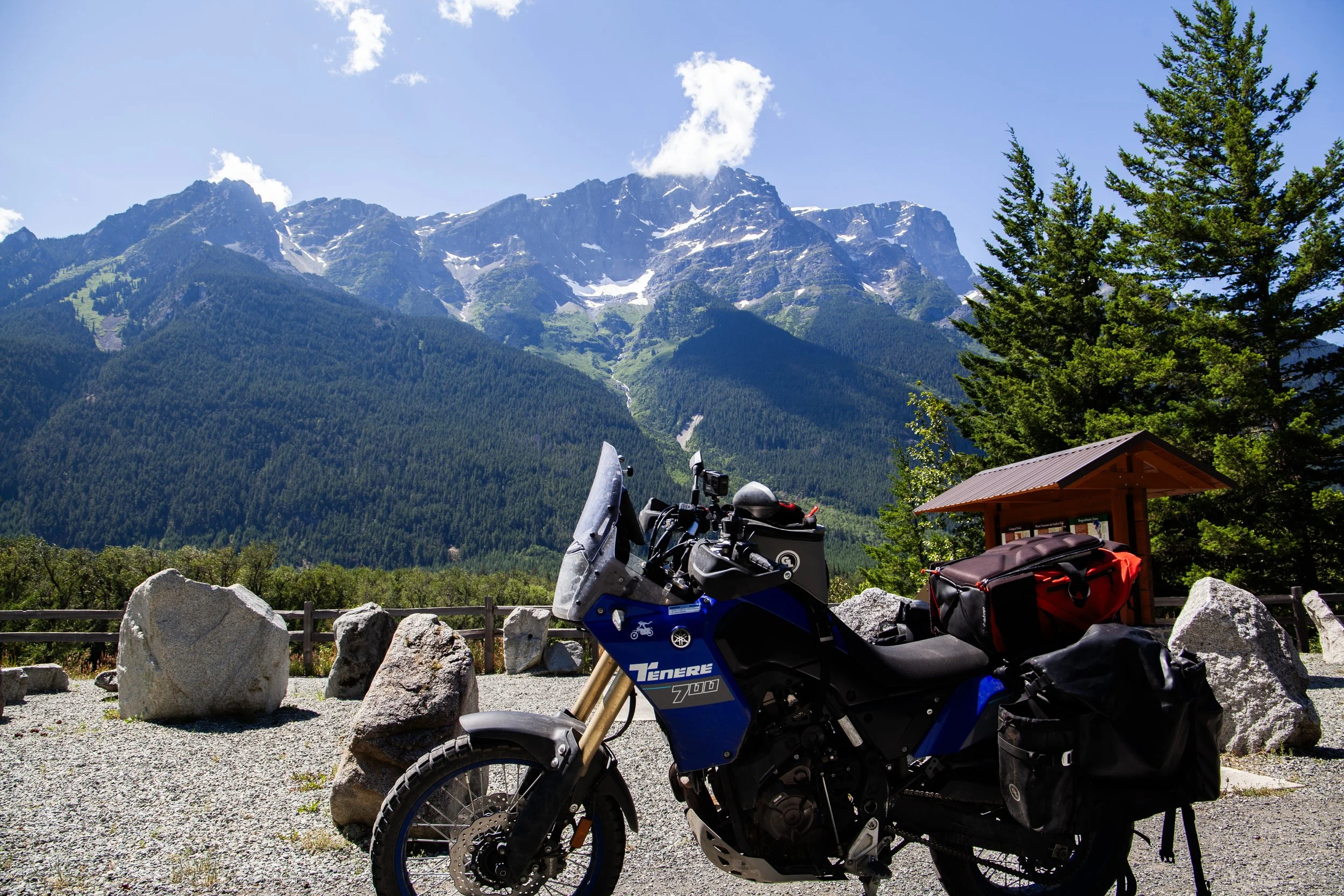 A blue adventure motorcycle parked on gravel with a mountain range and green trees in the background under a partly cloudy sky.