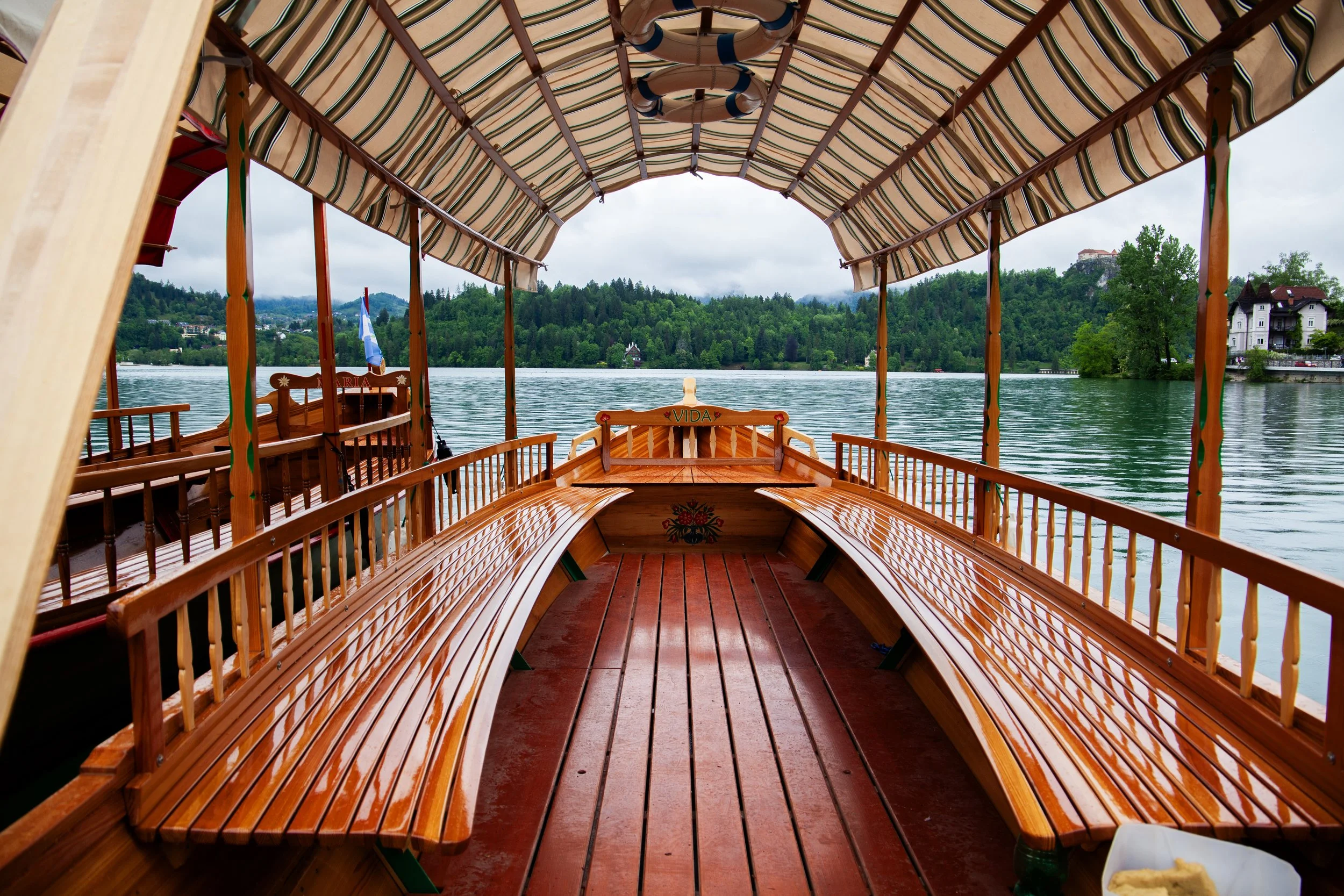 View from inside a wooden boat with curved benches and a striped canopy, cruising on a river with green trees and houses in the background.