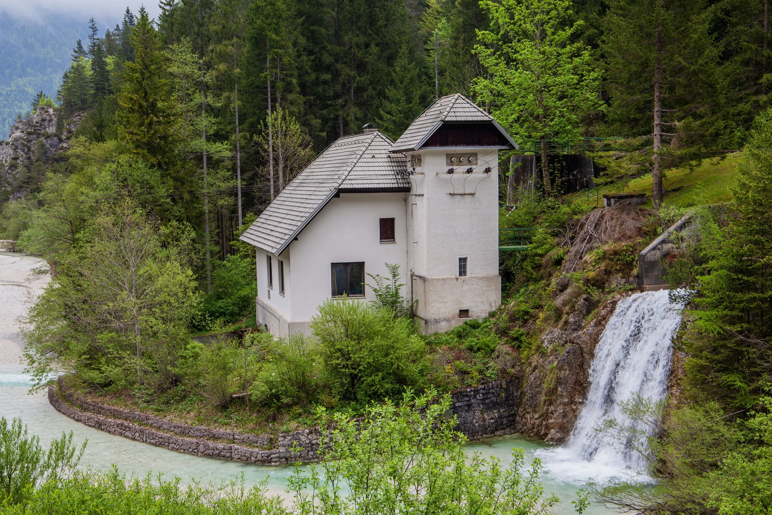 White house with multiple roofs situated on a narrow land surrounded by green trees and a small waterfall cascading into a river.