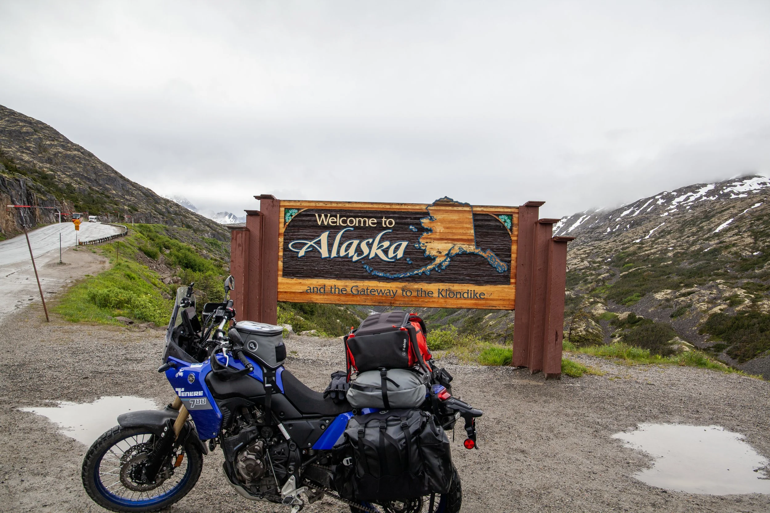 A motorcycle parked in front of a welcome sign that reads 'Welcome to Alaska and the Gateway to the Klondike.' The sign features a map outline of Alaska. In the background, there is a winding mountain road, some green vegetation, mountainous terrain, and cloudy skies.