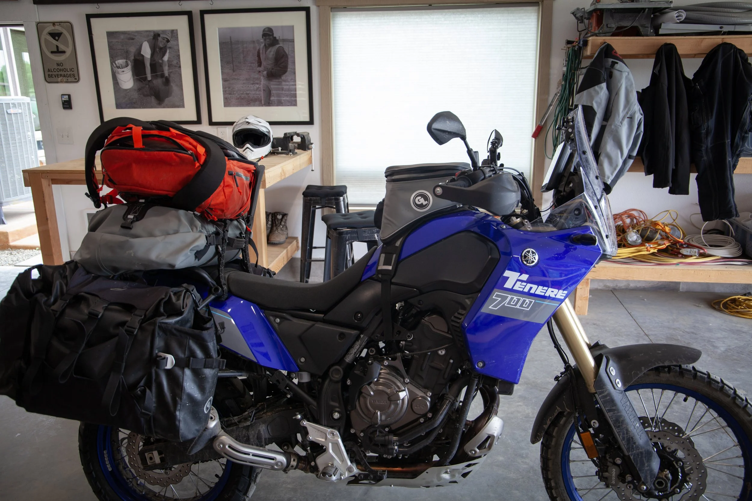 A Yamaha Tenere 700 motorcycle loaded with gear inside a garage. The bike has black saddlebags, a gray tank bag, and a red backpack on the rear. The garage has a workbench with pictures on the wall and some jackets hanging on a shelf.