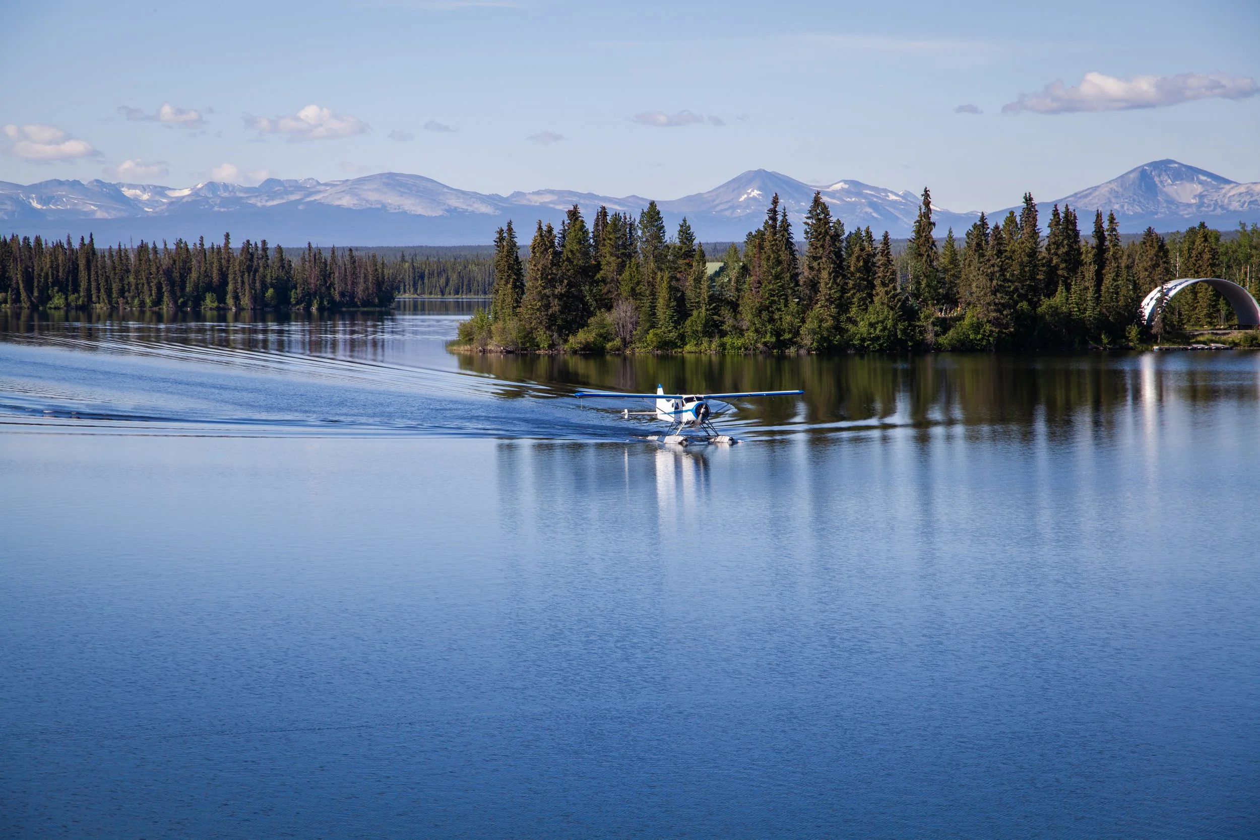 A small seaplane flying over a calm lake with trees and snow-capped mountains in the background.